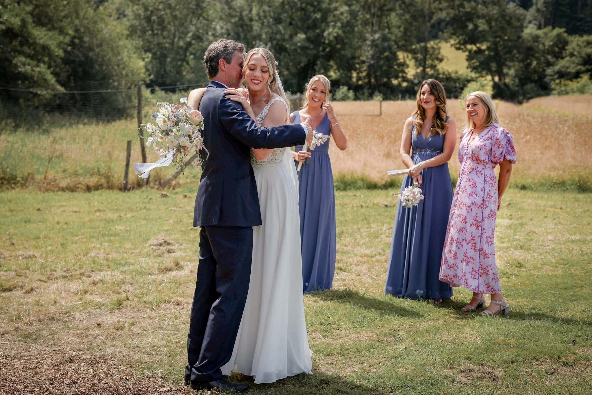 A wedding ceremony outdoors with a bride and groom embracing, surrounded by three bridesmaids in blue dresses and a woman in a pink patterned dress, on a grassy field with trees in the background.