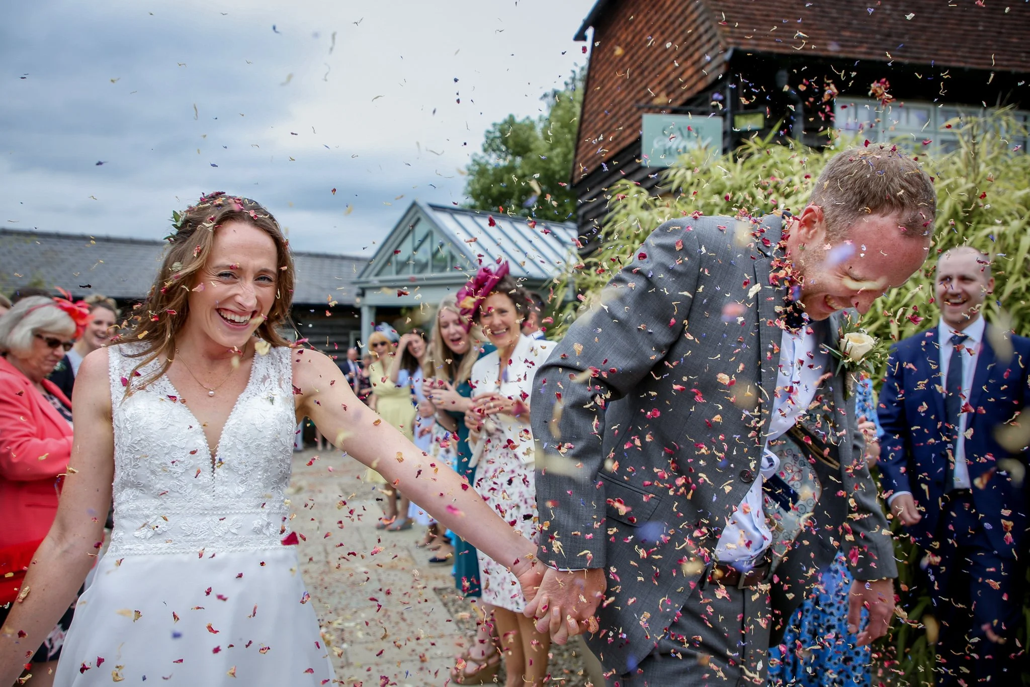 A newlywed couple holding hands and walking through a confetti celebration with friends and family during their outdoor wedding reception.