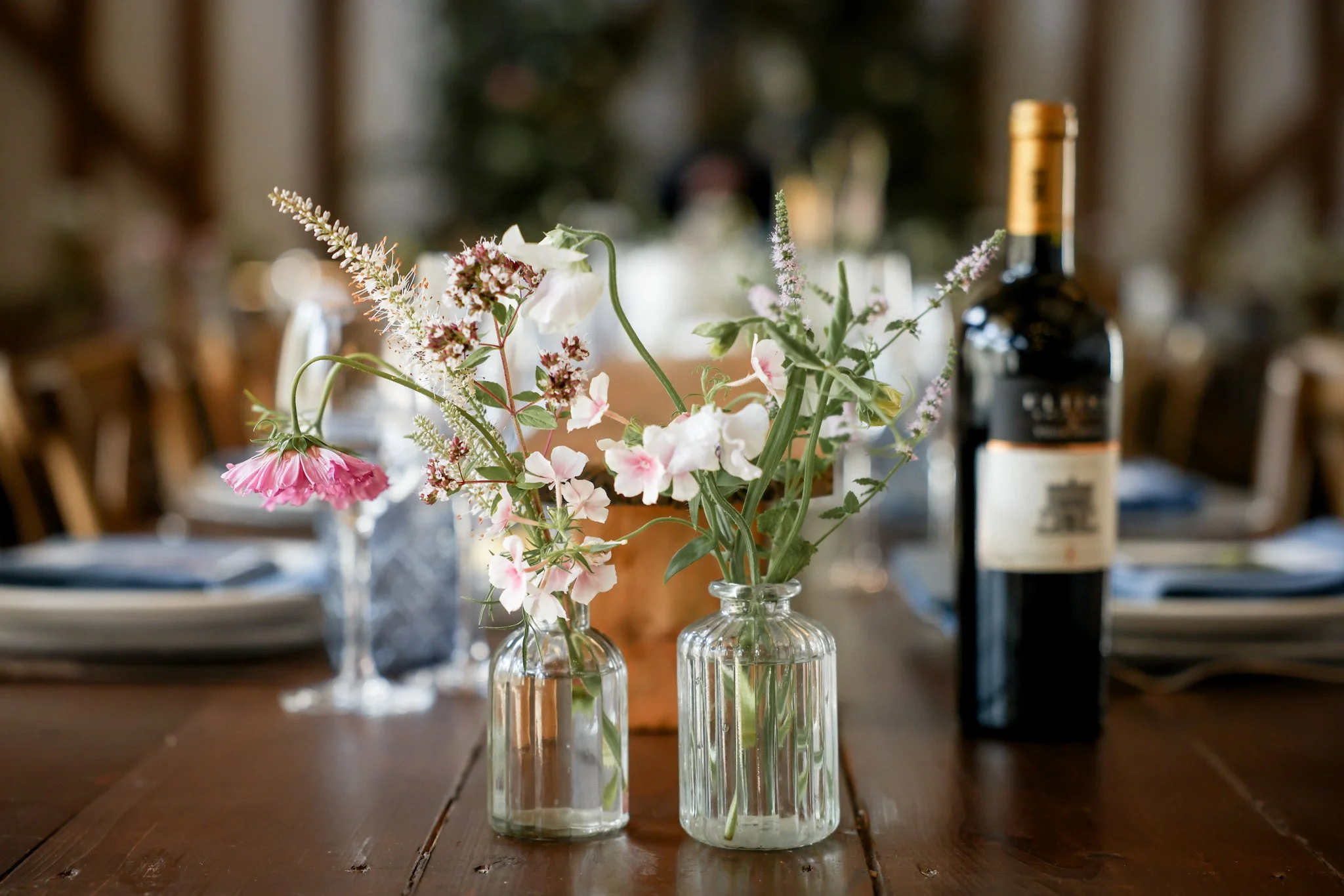 Three glass vases with assorted flowers, including pink, white, and purple blooms, are placed on a wooden table, with a wine bottle visible in the background.