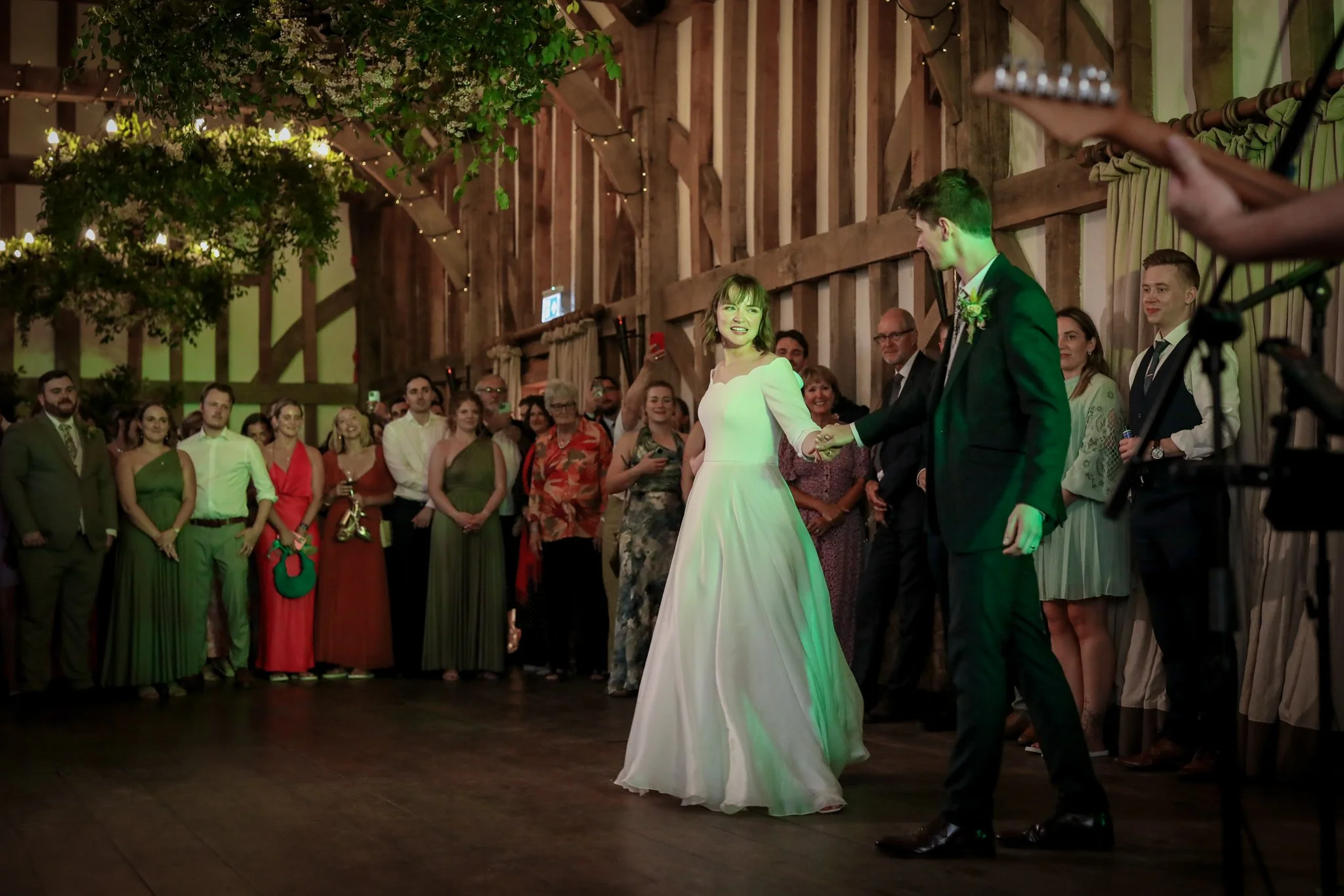 A newlywed couple is dancing during their wedding reception, surrounded by guests in a rustic indoor venue decorated with string lights and greenery.