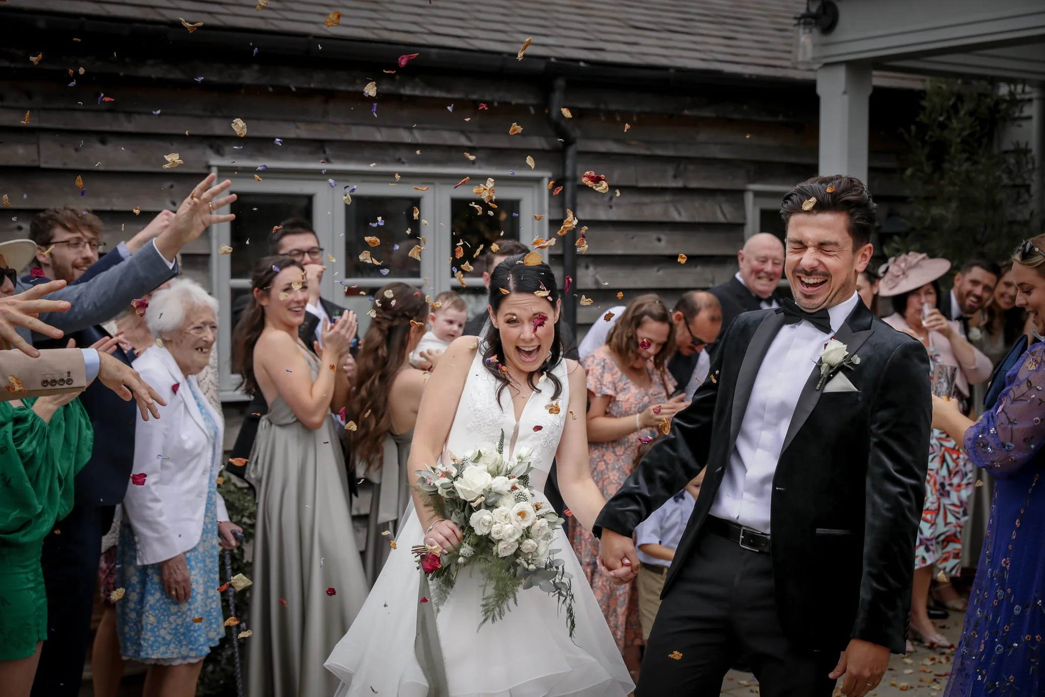 A wedding celebration with a bride and groom holding hands, surrounded by guests throwing flower petals, outside in front of a wooden house.