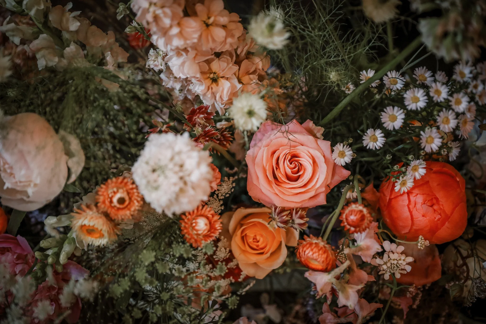 Close-up of various flowers including peach roses, white daisies, small orange flowers, and cream-colored blooms with green foliage.