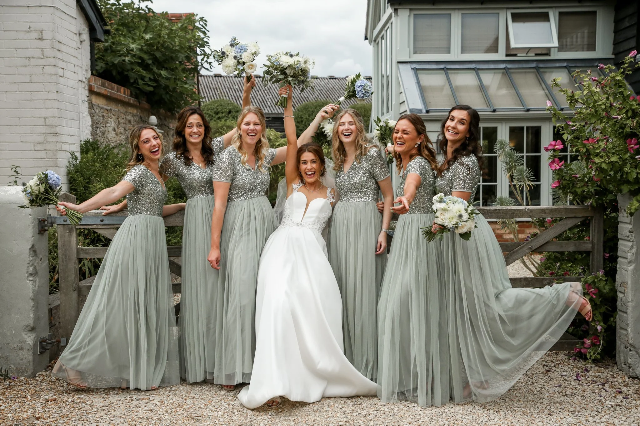 A bride and six bridesmaids are celebrating outdoors near a garden gate. The bridesmaids are wearing matching silver sequined tops and long, light green skirts, holding bouquets, and raising their arms in celebration. The bride is in a white gown wit