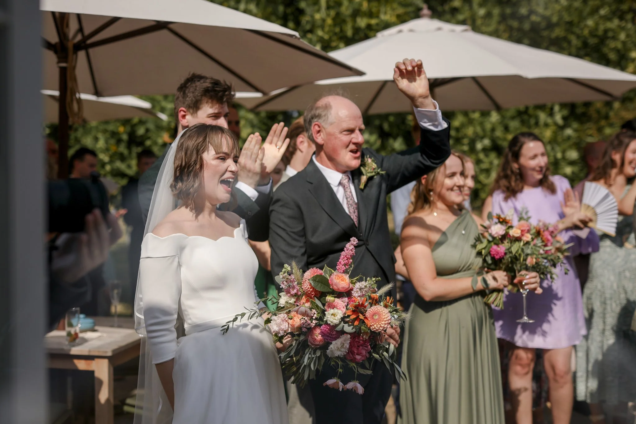 Group of people, including a bride and groom, participating in a wedding celebration outdoors, with guests holding flowers and smiling under umbrellas.