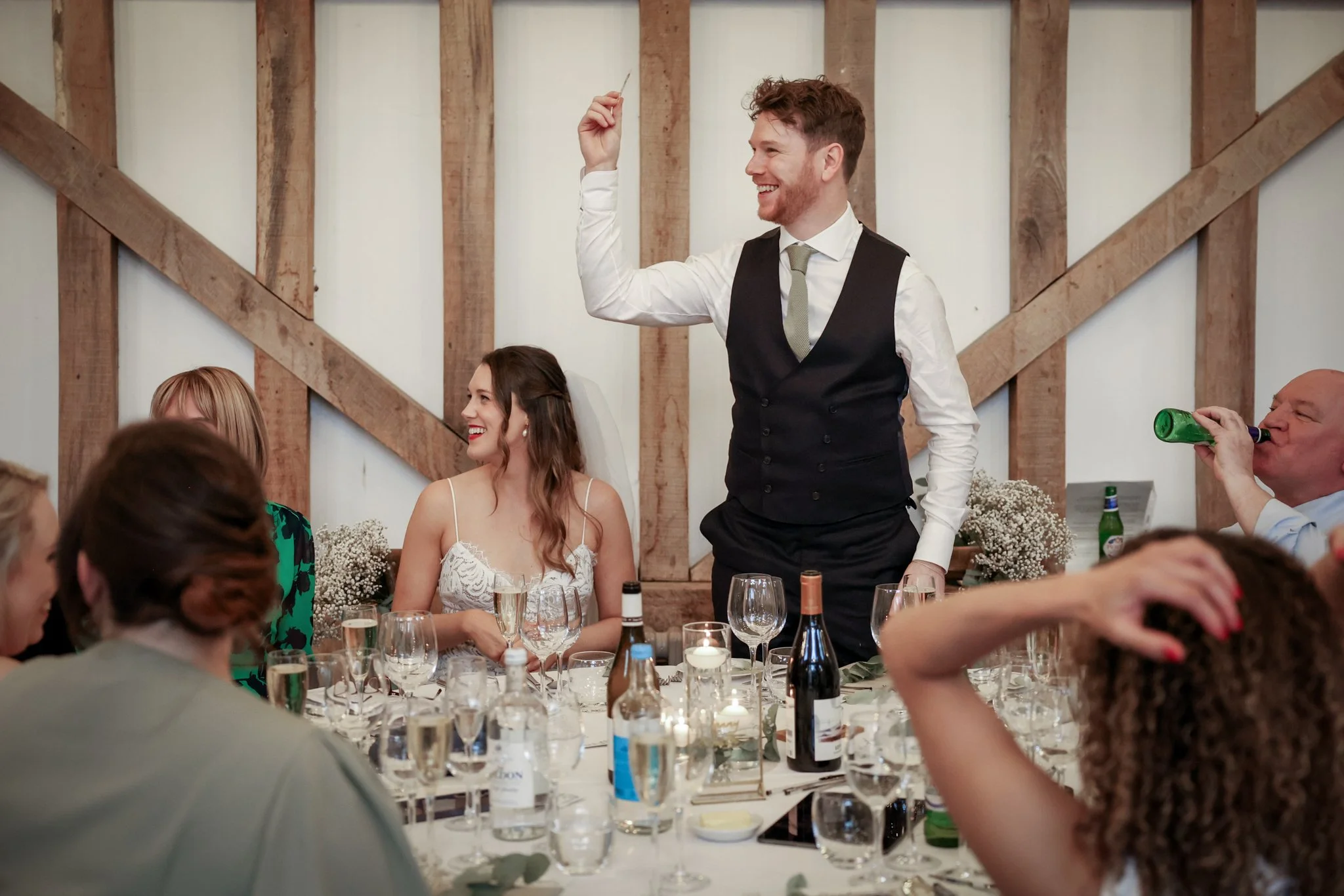 A man dressed in formal wedding attire, including a vest and tie, smiling and raising a glass or a utensil at a wedding reception. He is surrounded by seated guests, a bride with a veil, and a rustic wooden interior background.