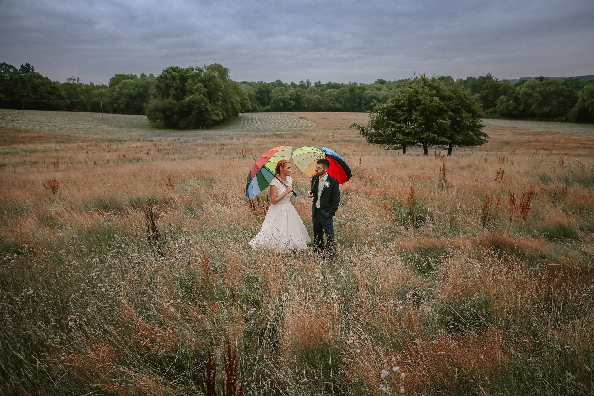 A bride and groom standing in a field holding a multicolored umbrella, with trees and cloudy sky in the background.