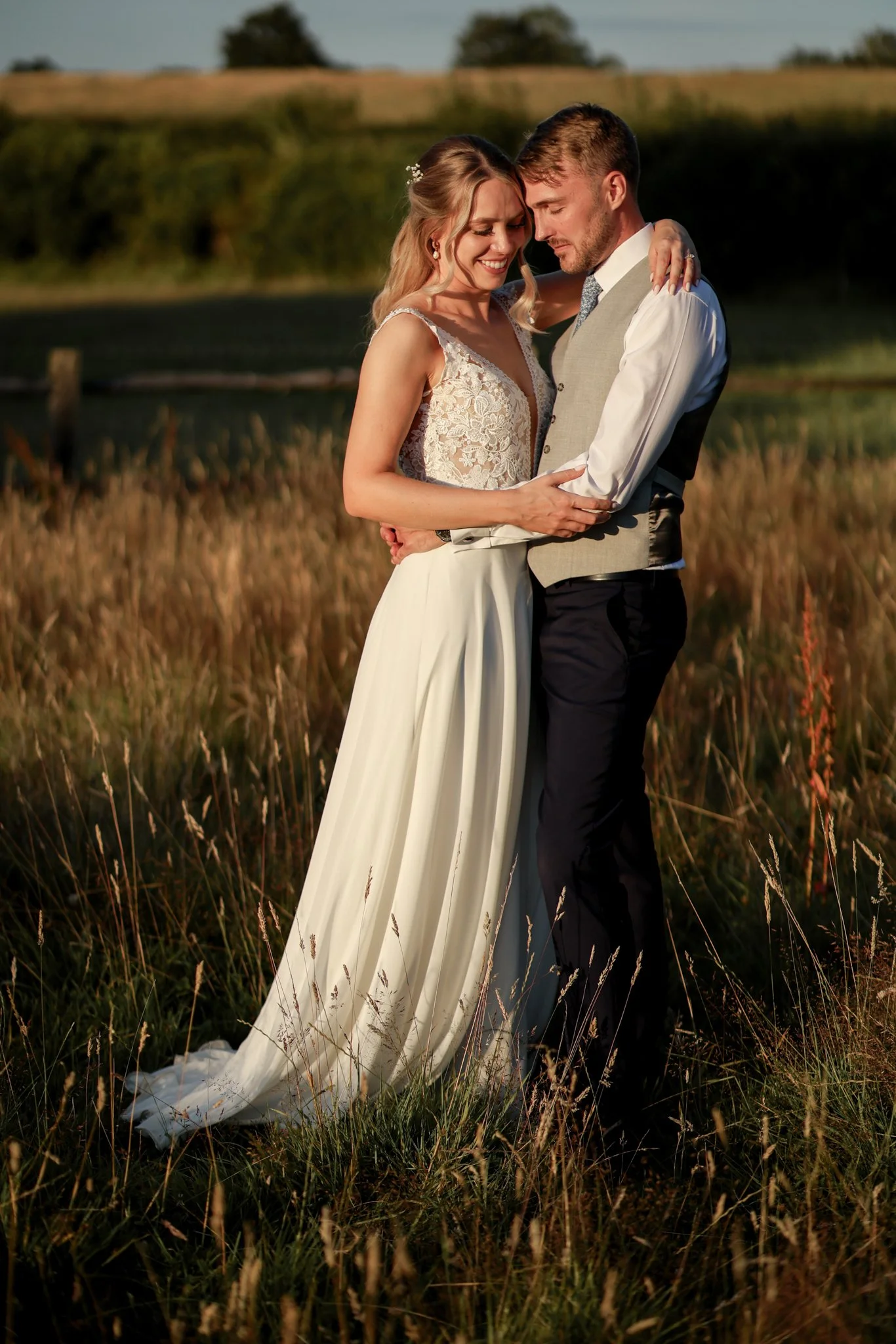 A bride and groom embracing in a field during sunset, dressed in wedding attire, smiling softly with eyes closed.