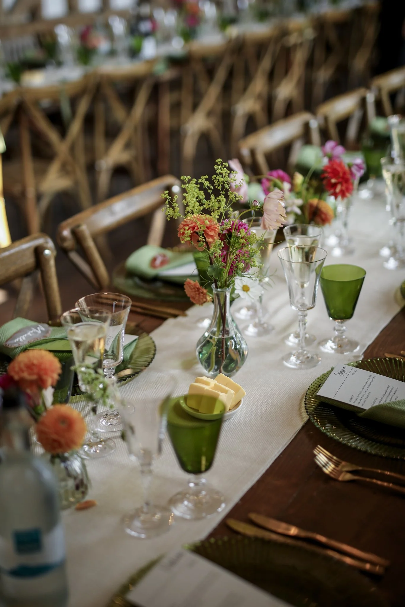 A decorated dining table set for a special event with colorful flowers in a vase, glasses, butter, and place settings, with wooden chairs in the background.