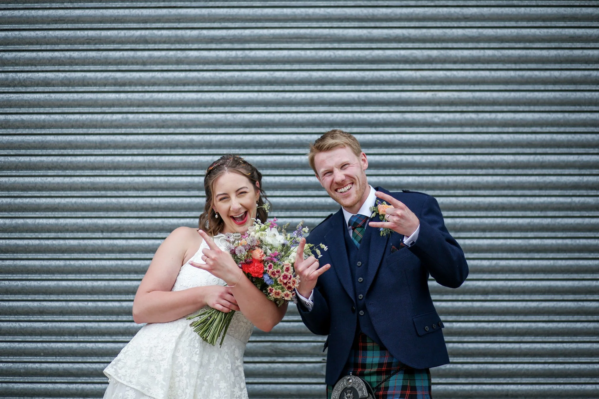 A happy couple in wedding attire posing in front of a metal rolling door, with the bride holding a bouquet of colorful flowers and both making rock on hand gestures.