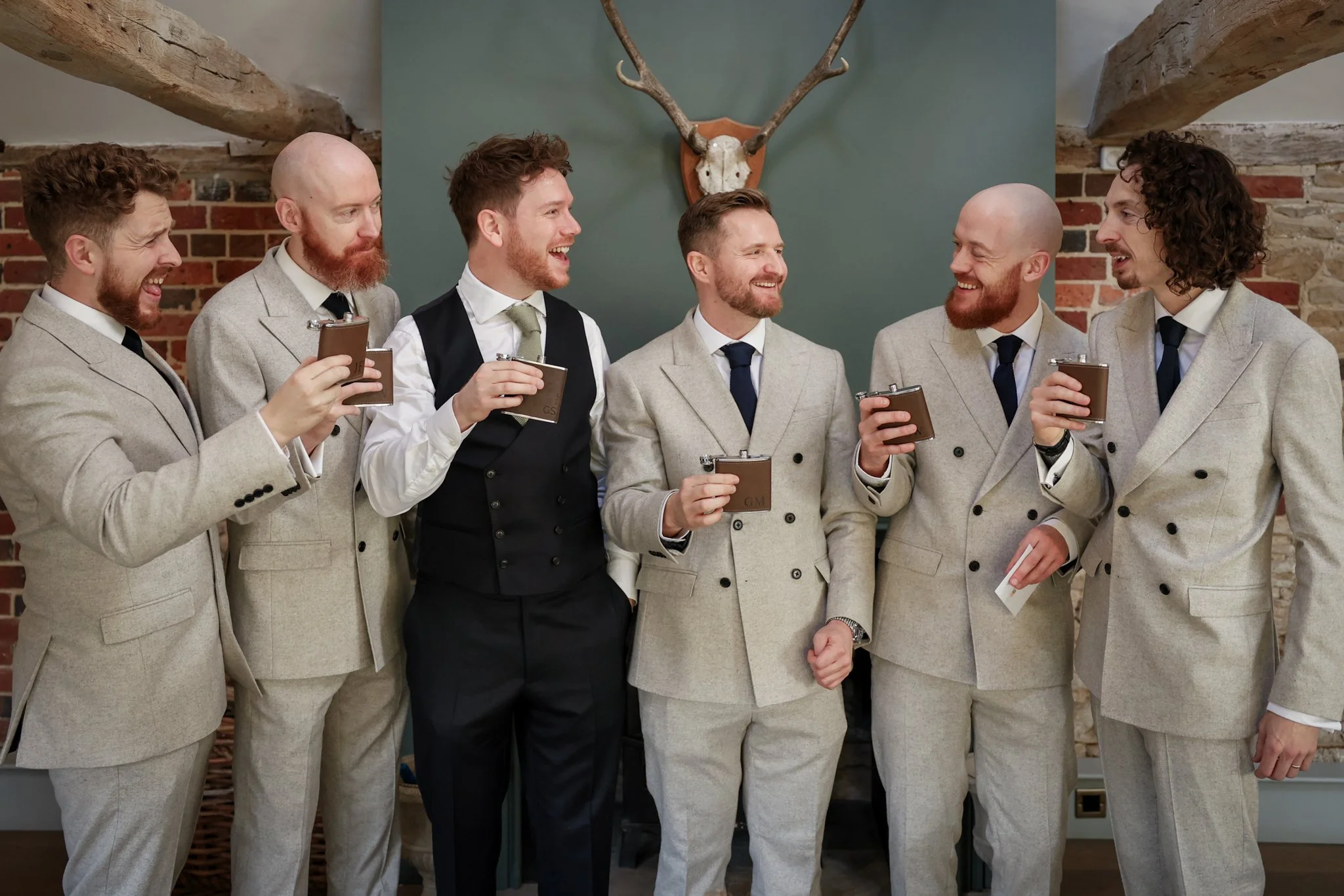 A group of six men in suits and ties, smiling and holding drinks, gathered in a rustic room with brick walls and a deer antler wall decoration behind them.
