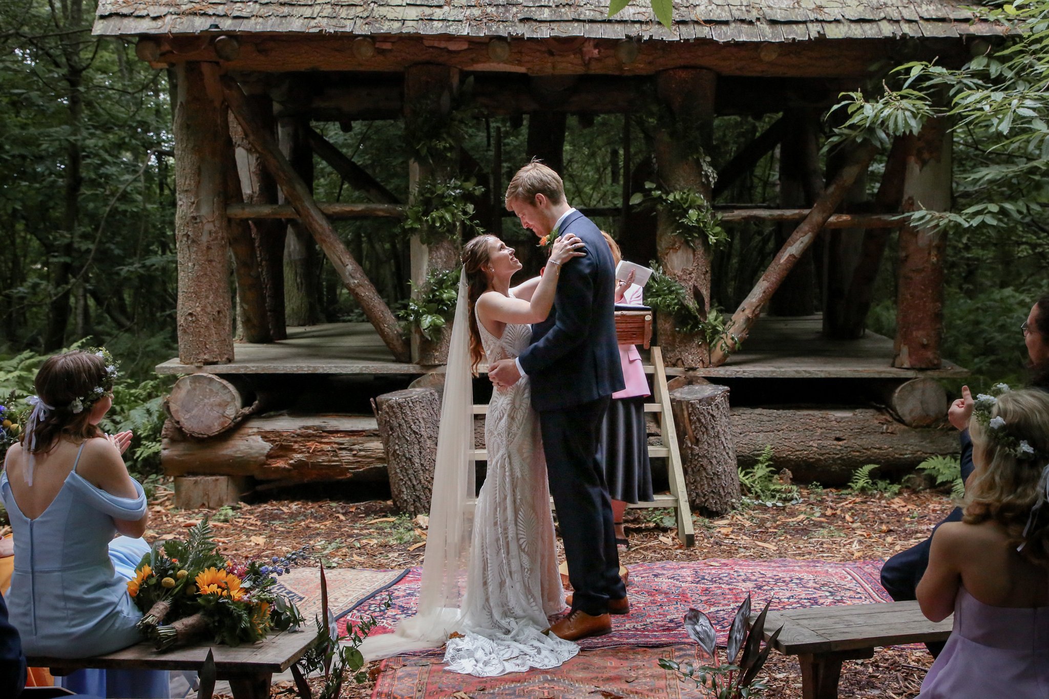 A bride and groom exchange vows during an outdoor wedding ceremony in a wooded area, with guests seated around them, and a rustic wooden structure in the background.