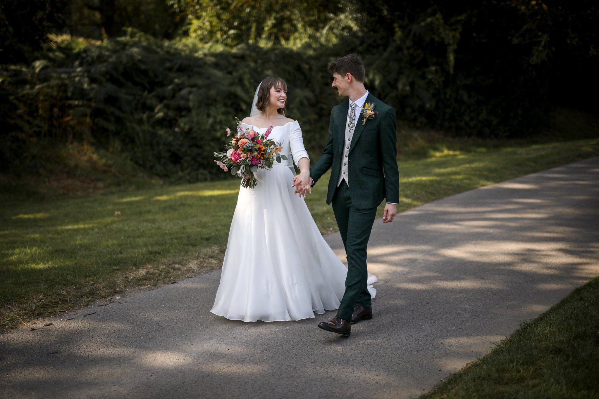 A bride and groom holding hands and smiling at each other in an outdoor setting with greenery.