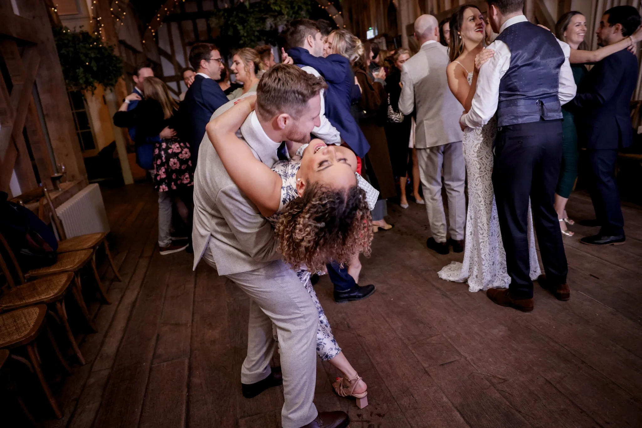 People dancing and enjoying themselves at a lively indoor party or wedding reception with rustic decor and string lights.