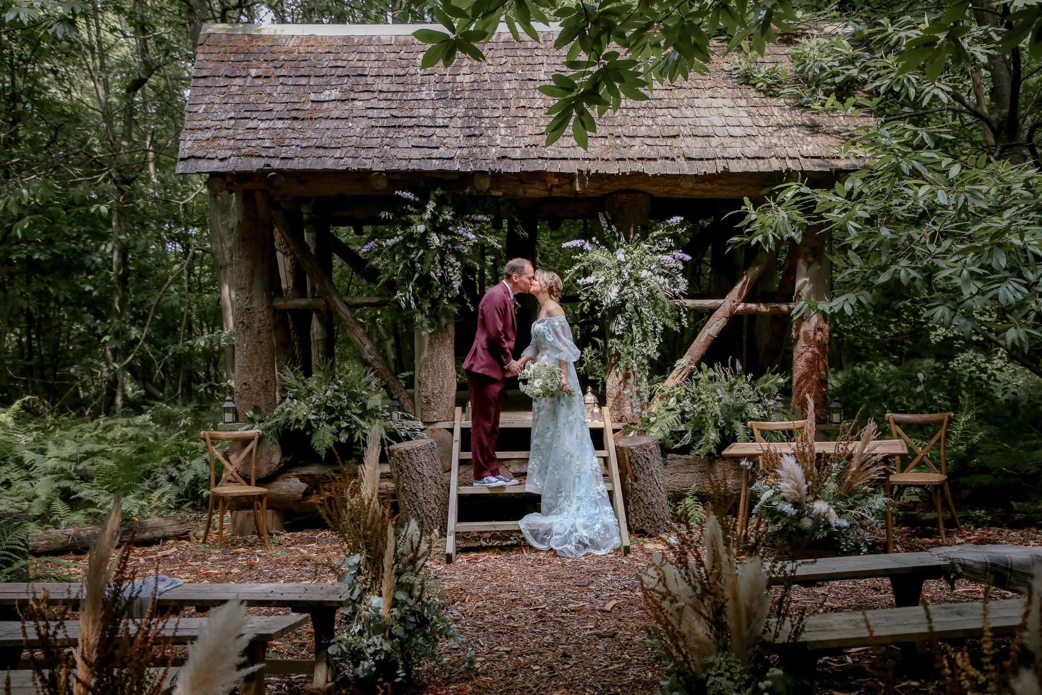 A bride and groom sharing a kiss at an outdoor wedding ceremony in a forest setting on a wooden stage decorated with flowers and greenery.