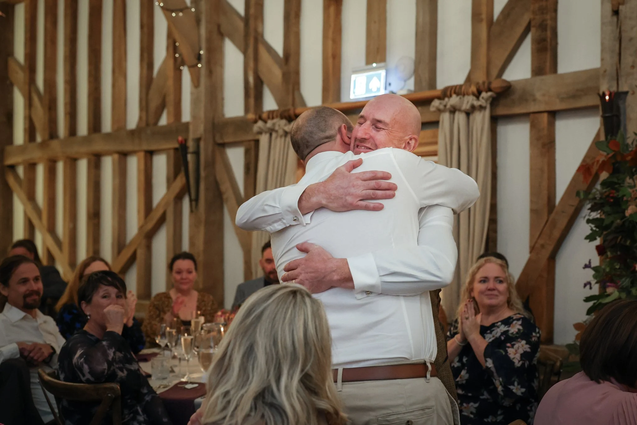 Two men hug at a wedding reception, surrounded by seated guests in a rustic barn setting with wood beams and string lights.