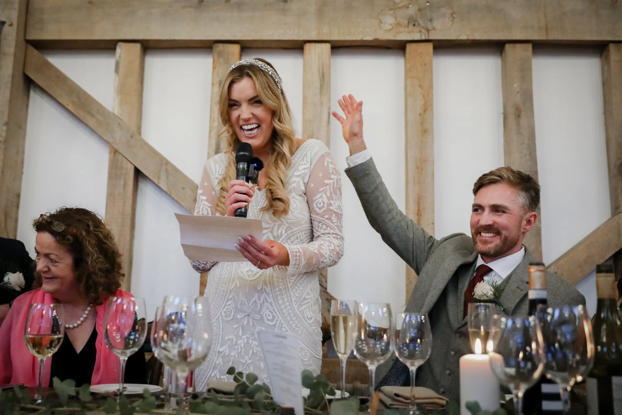 A woman in a white wedding dress wearing a tiara is speaking into a microphone at a wedding reception. A man next to her is smiling and raising his hand. Other guests are seated at a table with wine glasses and candles.