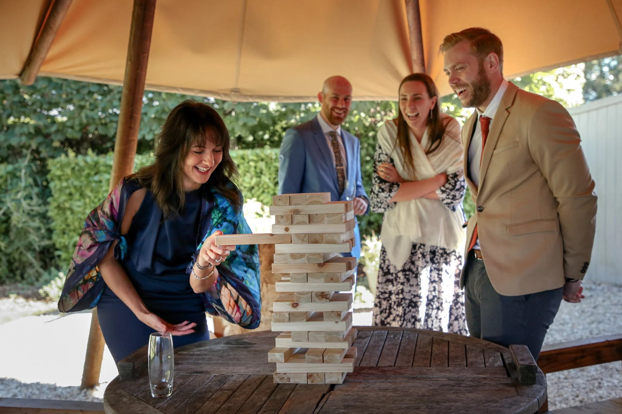 A group of five people playing a large outdoor Jenga game under a canopy with greenery in the background. One woman in a colorful jacket is taking her turn, with four others watching and laughing.