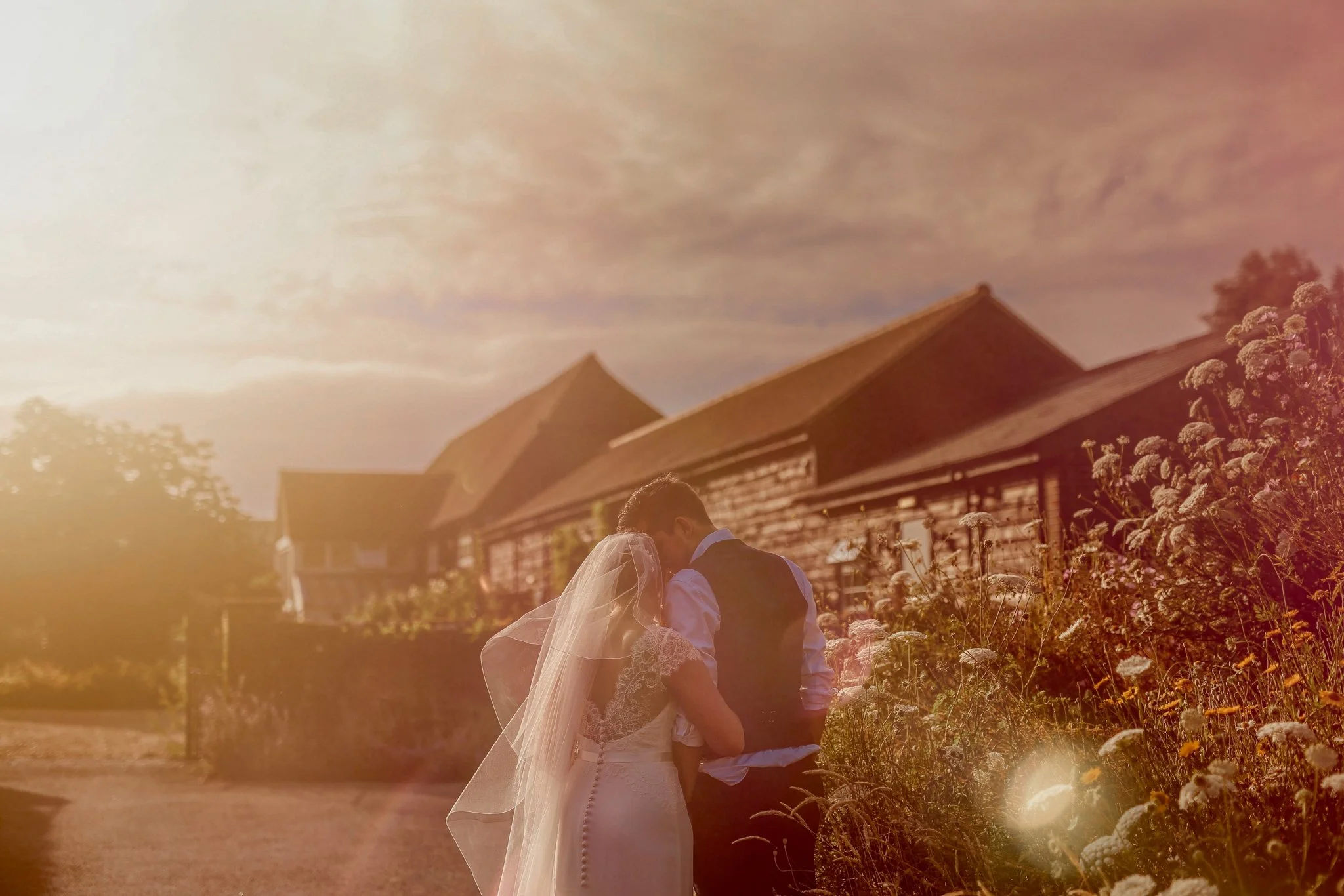 A bride and groom share an intimate moment in a field of tall flowers during sunset, with rustic buildings in the background.
