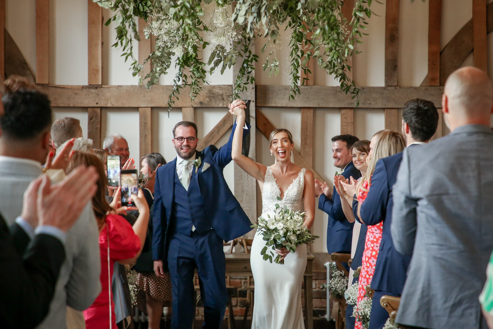 A bride and groom holding hands and smiling while walking down the aisle during a wedding ceremony, surrounded by seated guests in a rustic venue with wooden beams and hanging greenery.