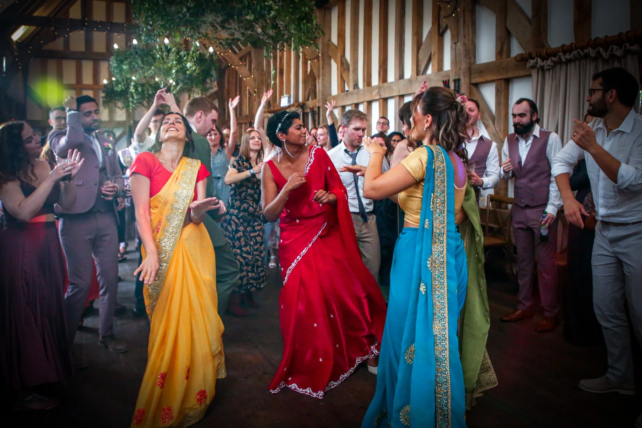 People dancing at a wedding reception, including women in colorful traditional dresses and men in formal attire, inside a rustic barn with fairy lights and greenery.
