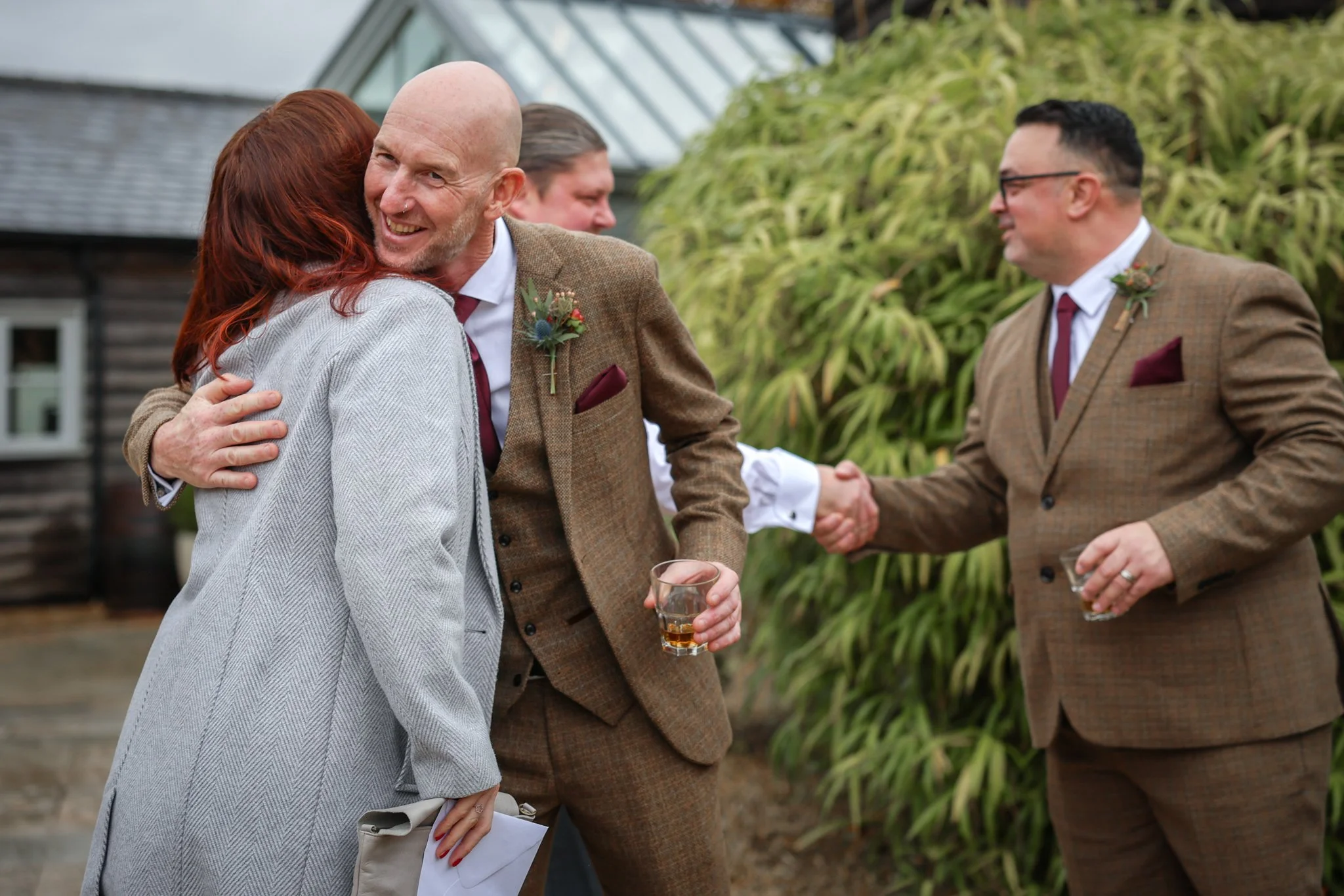 People at a social gathering outdoors, smiling, hugging, and shaking hands, with some holding drinks.
