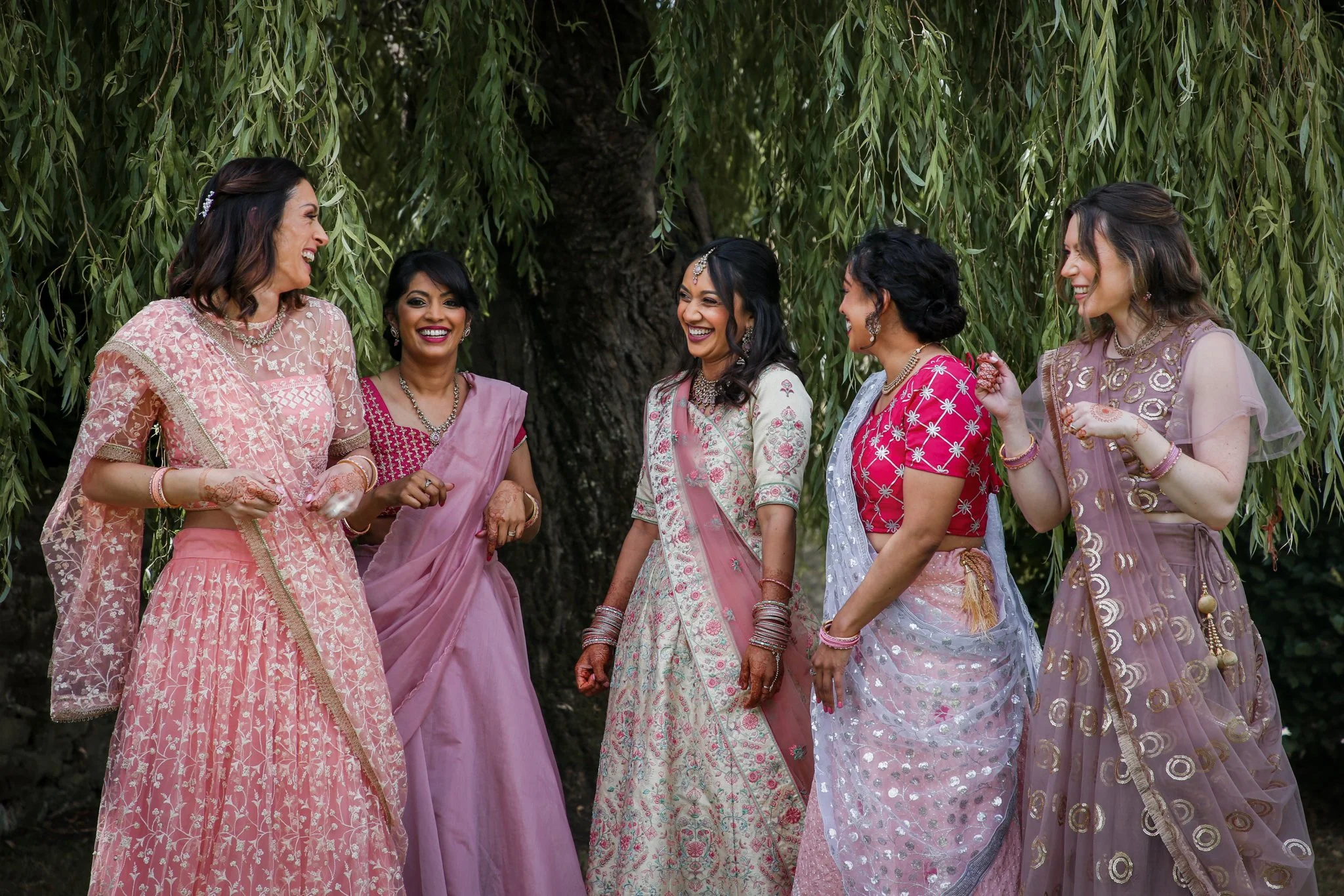 Group of women wearing traditional Indian attire, standing outdoors under a large tree, smiling and chatting.