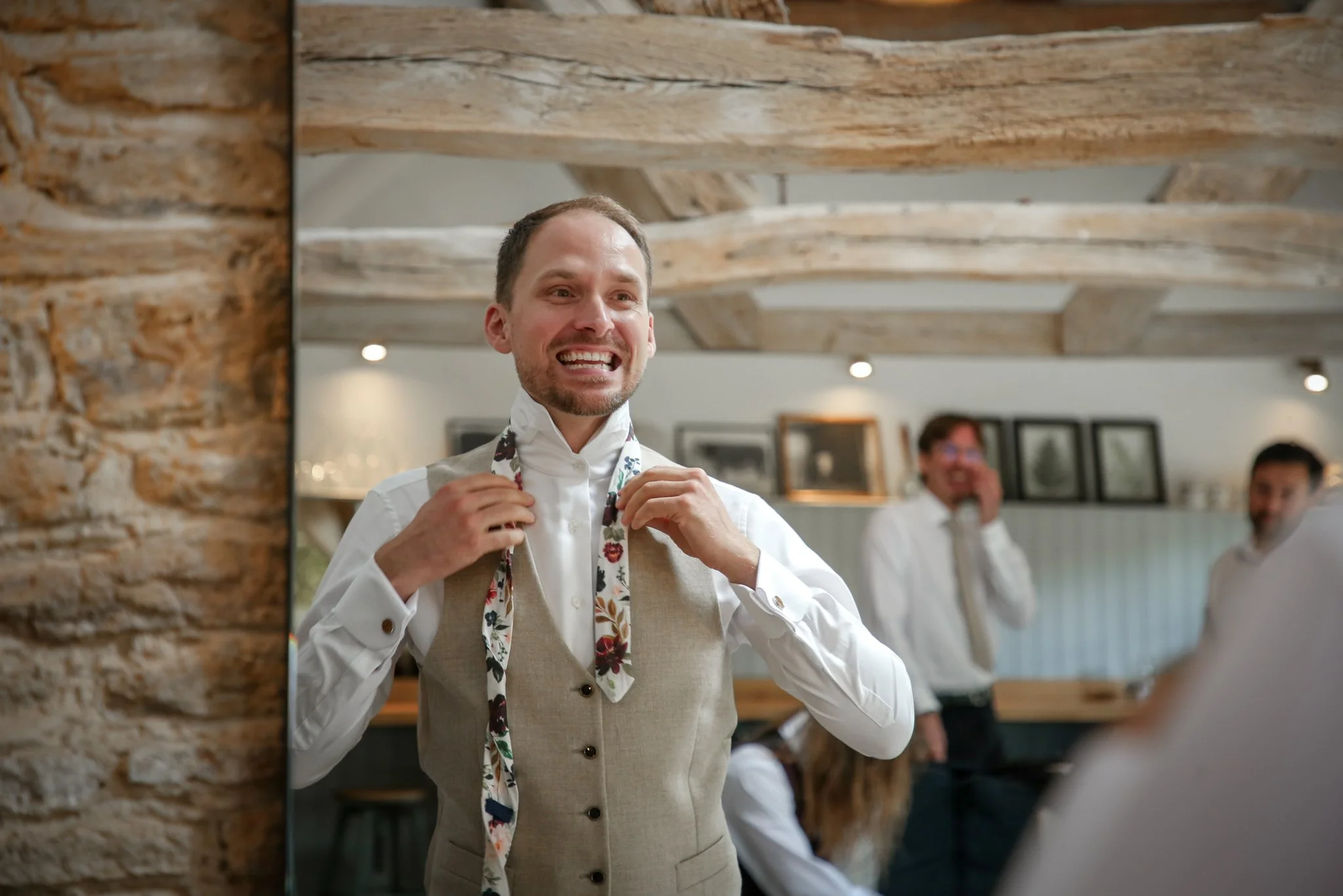 A smiling man adjusting his floral apron in a restaurant setting, with exposed wooden beams on the ceiling and other staff members in the background.