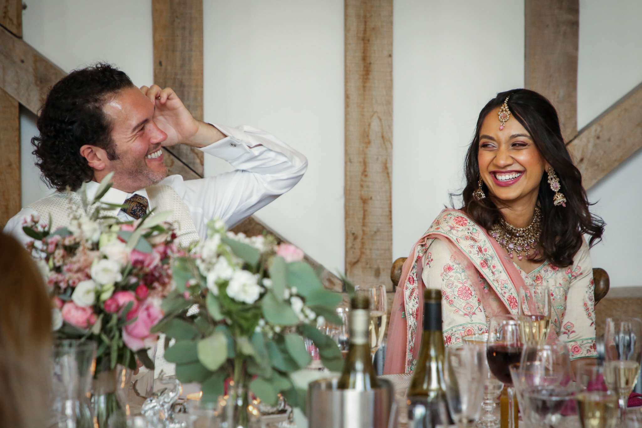 A happy man in a white shirt and vest, and a woman in traditional Indian attire with jewelry, are smiling and looking at each other at a decorated event table with flowers and drinks.