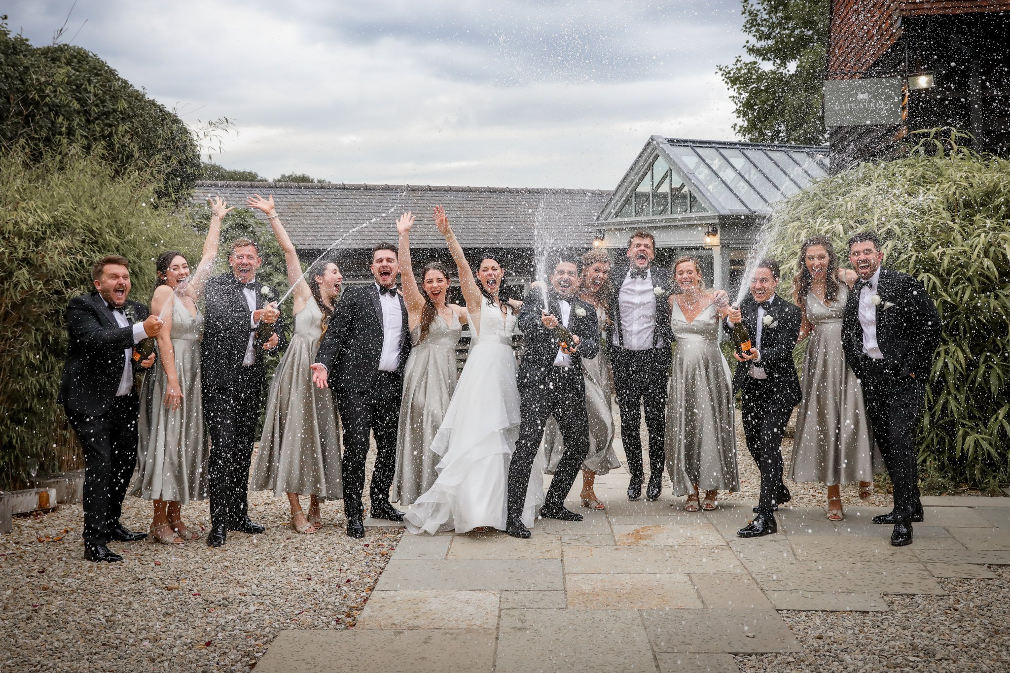 Wedding party celebrating outdoors, spraying champagne, on a cloudy day.