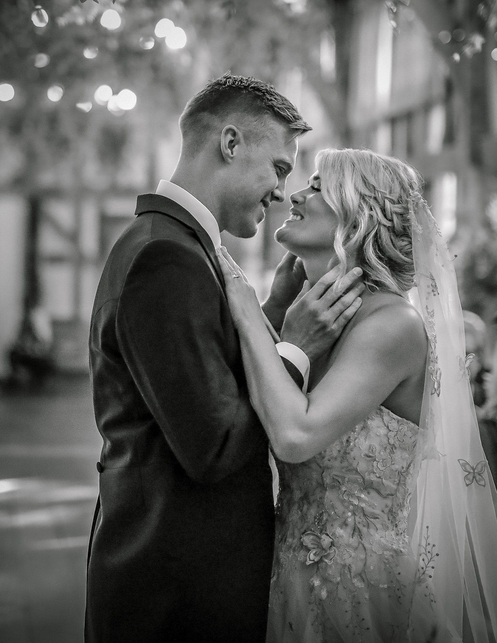A black and white photo of a couple dancing closely at their wedding, with the man wearing a tuxedo and the woman wearing a lace wedding dress with a veil, in an indoor setting.