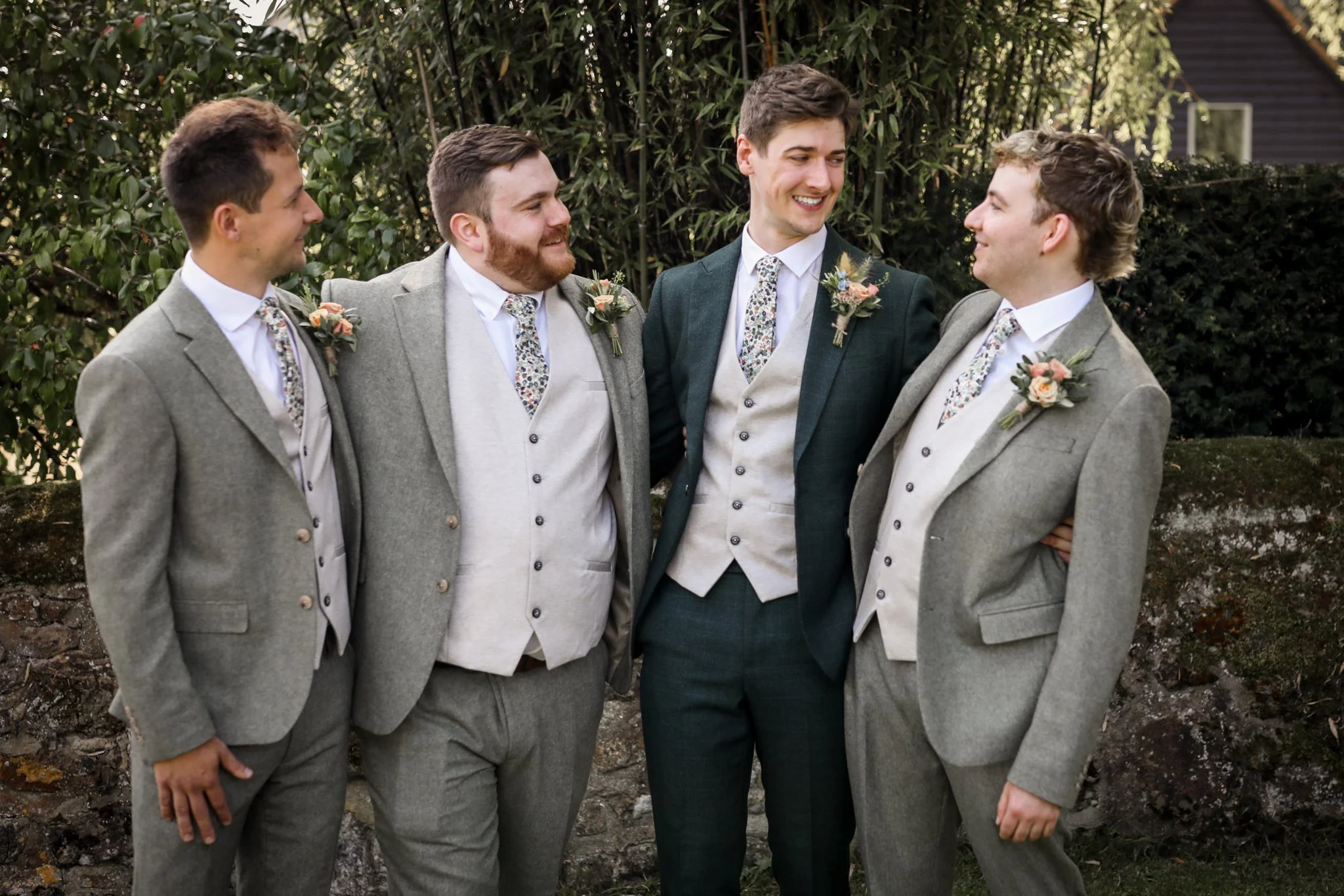Five men dressed in suits and flower boutonnieres standing outdoors, smiling and looking at each other, with greenery in the background.