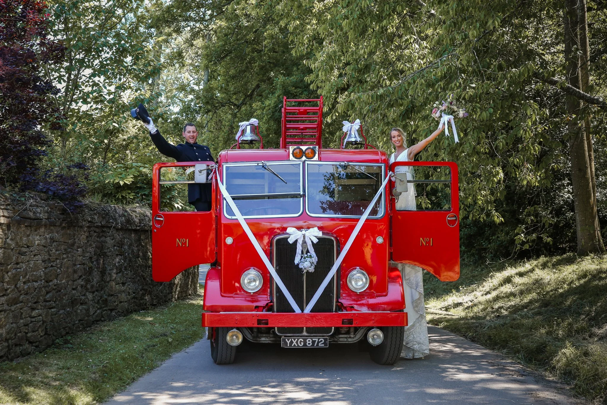 A bride and groom standing on a red fire truck decorated with white ribbons and ribbons, celebrating outdoors surrounded by green trees.