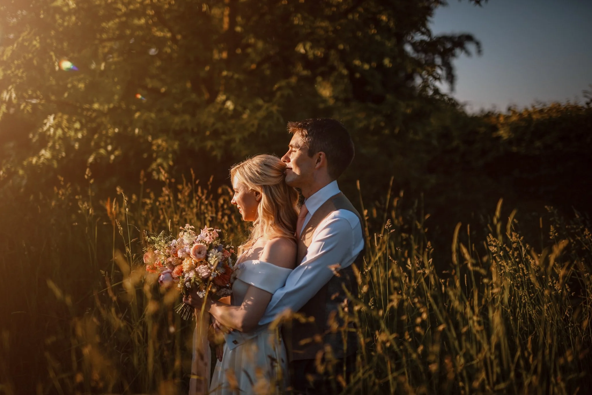 A happy couple in wedding attire embracing outdoors at sunset, with trees and tall grass in the background, the bride holding a bouquet of pink and white flowers.