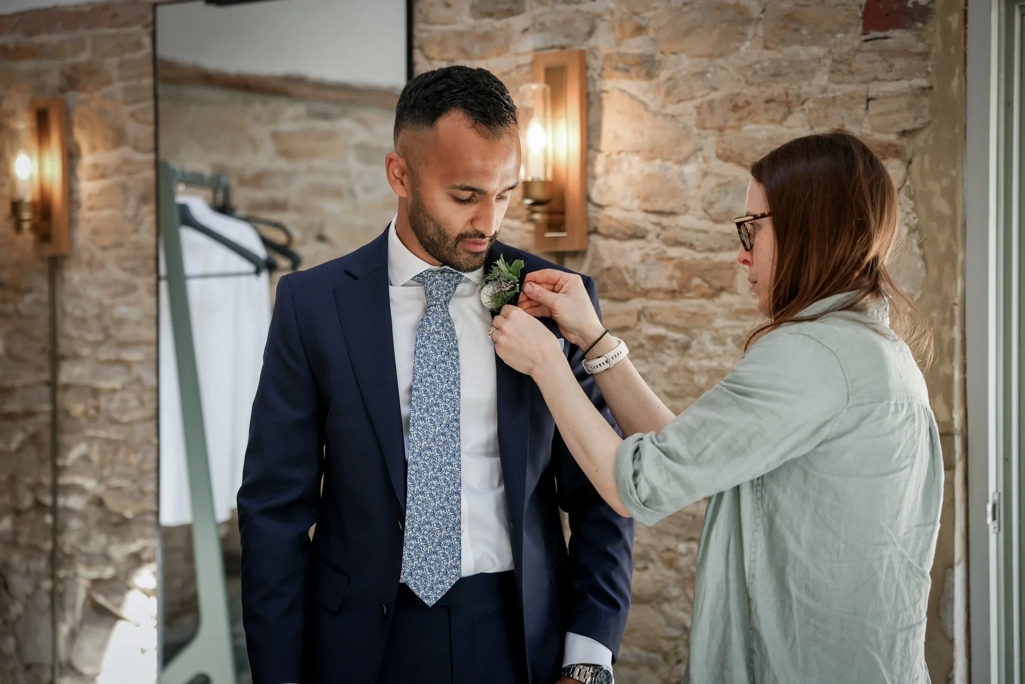 A woman pins a boutonniere onto a man in a navy suit, who is looking down in front of a rustic stone wall and a mirror.