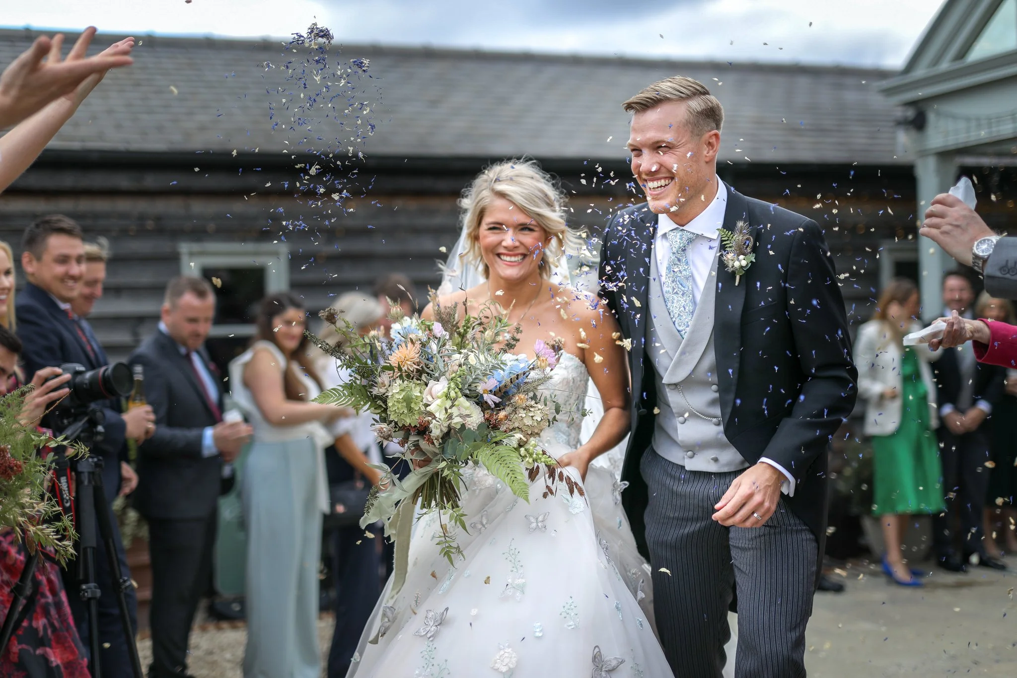 A newly married couple is celebrating with friends and family, throwing confetti during their outdoor wedding reception. The bride is smiling, holding a large bouquet of flowers, while the groom is dressed in a suit with a patterned tie and boutonnie