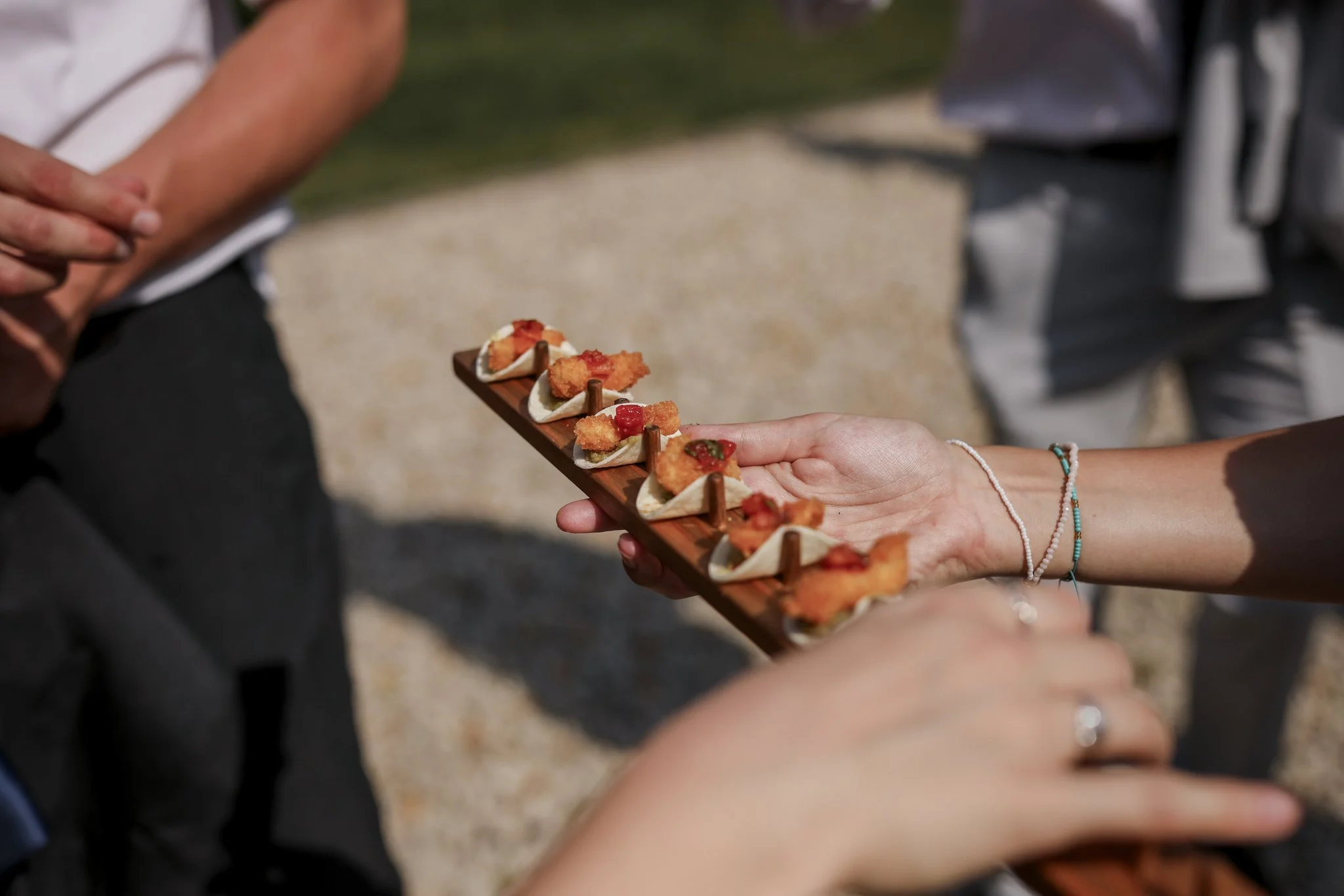 Hands holding a wooden tray with small appetizers, outdoors on a sunny day.