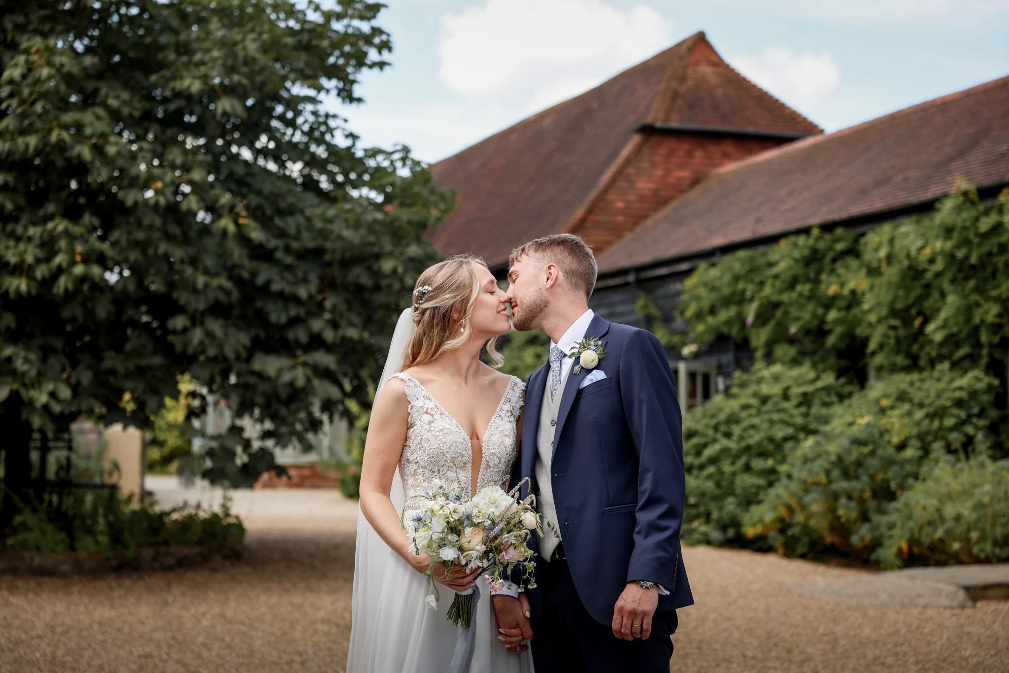 A bride and groom standing close, about to kiss, outdoors near a building with a sloped roof and green trees.