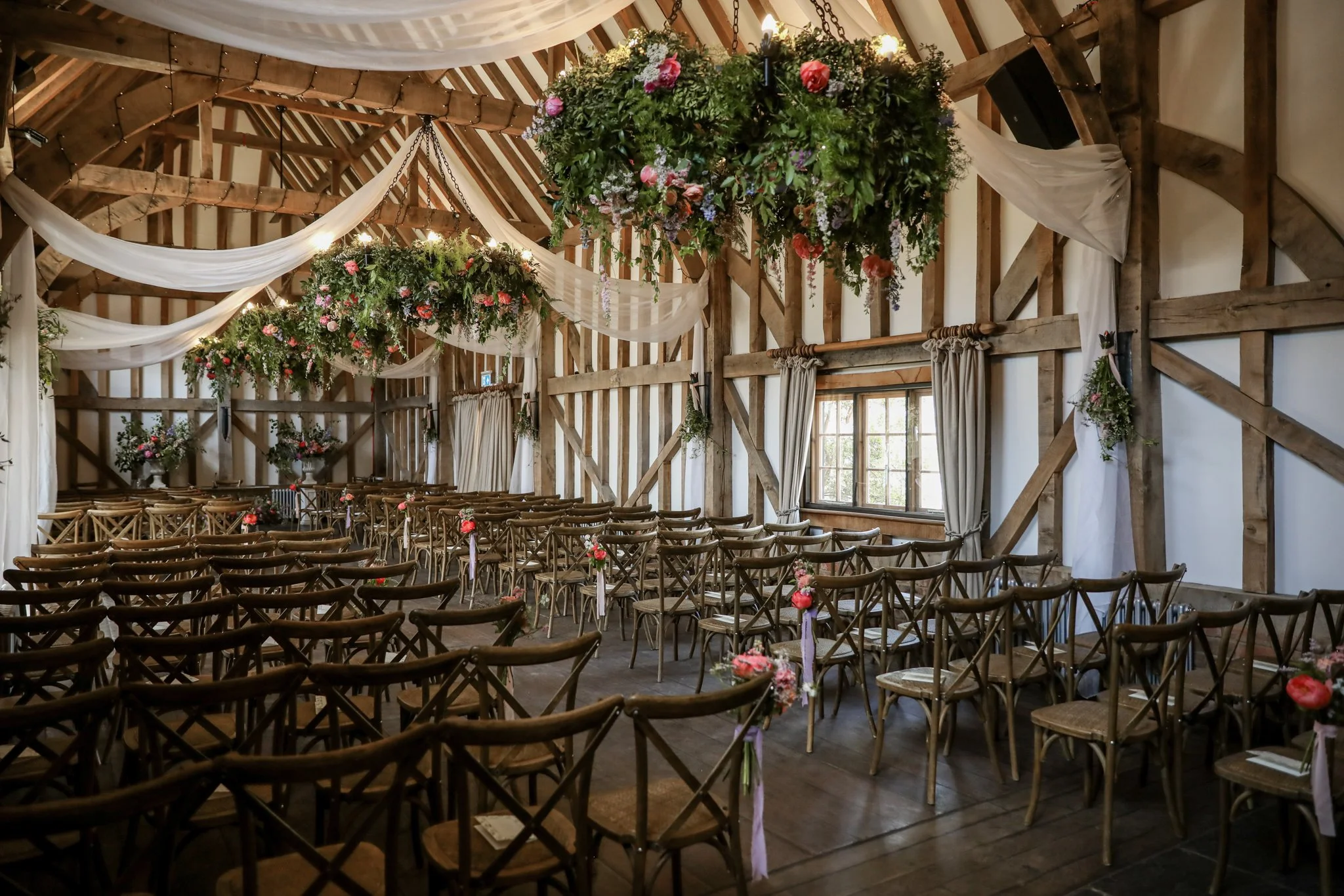 Interior of a rustic barn decorated for a wedding or special event, with wooden chairs arranged facing a decorated altar, large hanging floral arrangements, and white drapery adorning the ceiling.