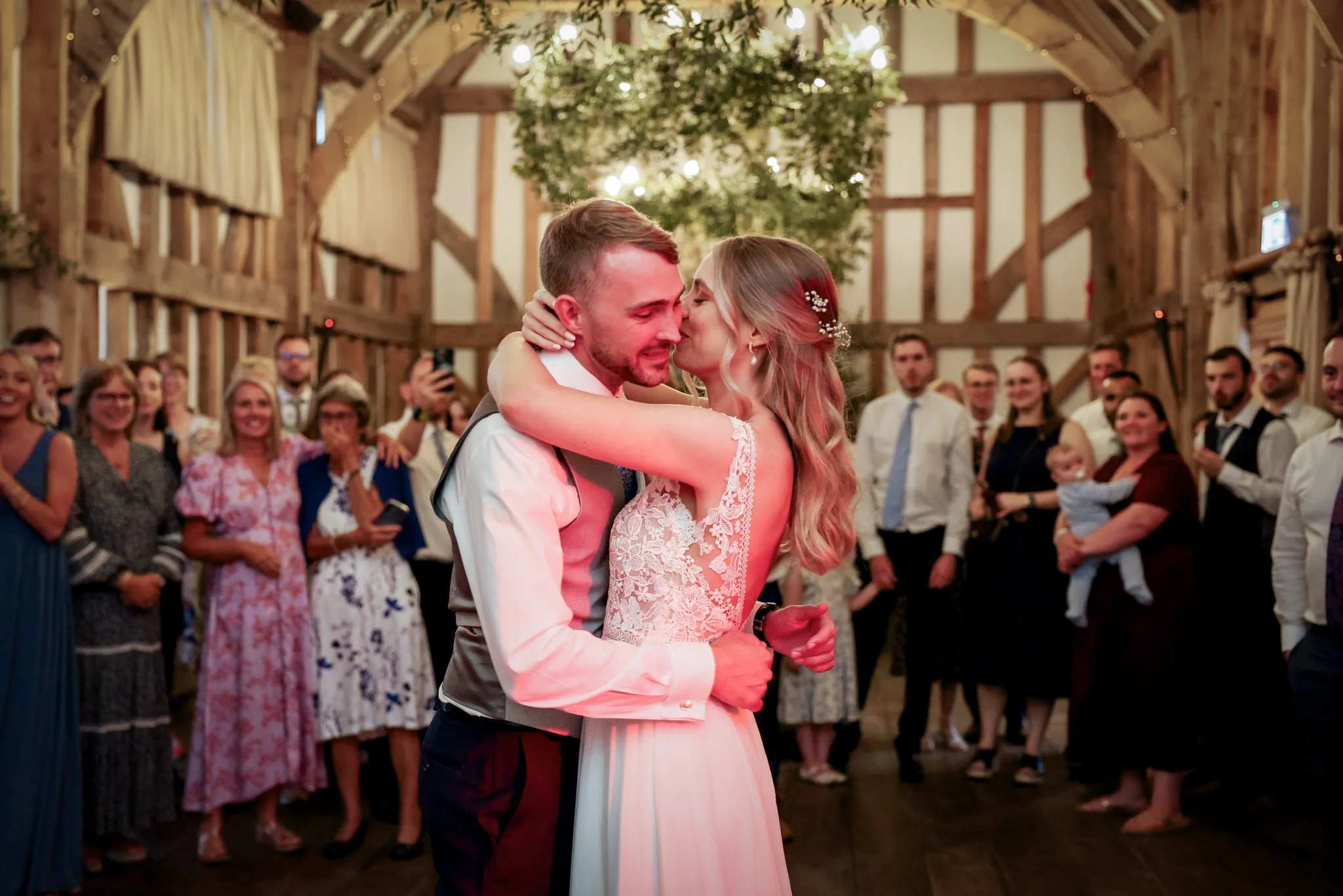 A bride and groom sharing a dance at a wedding reception. The bride wears a lace wedding gown, and the groom is dressed in a suit with a vest. They are surrounded by guests in a rustic barn setting with wooden beams and string lights.