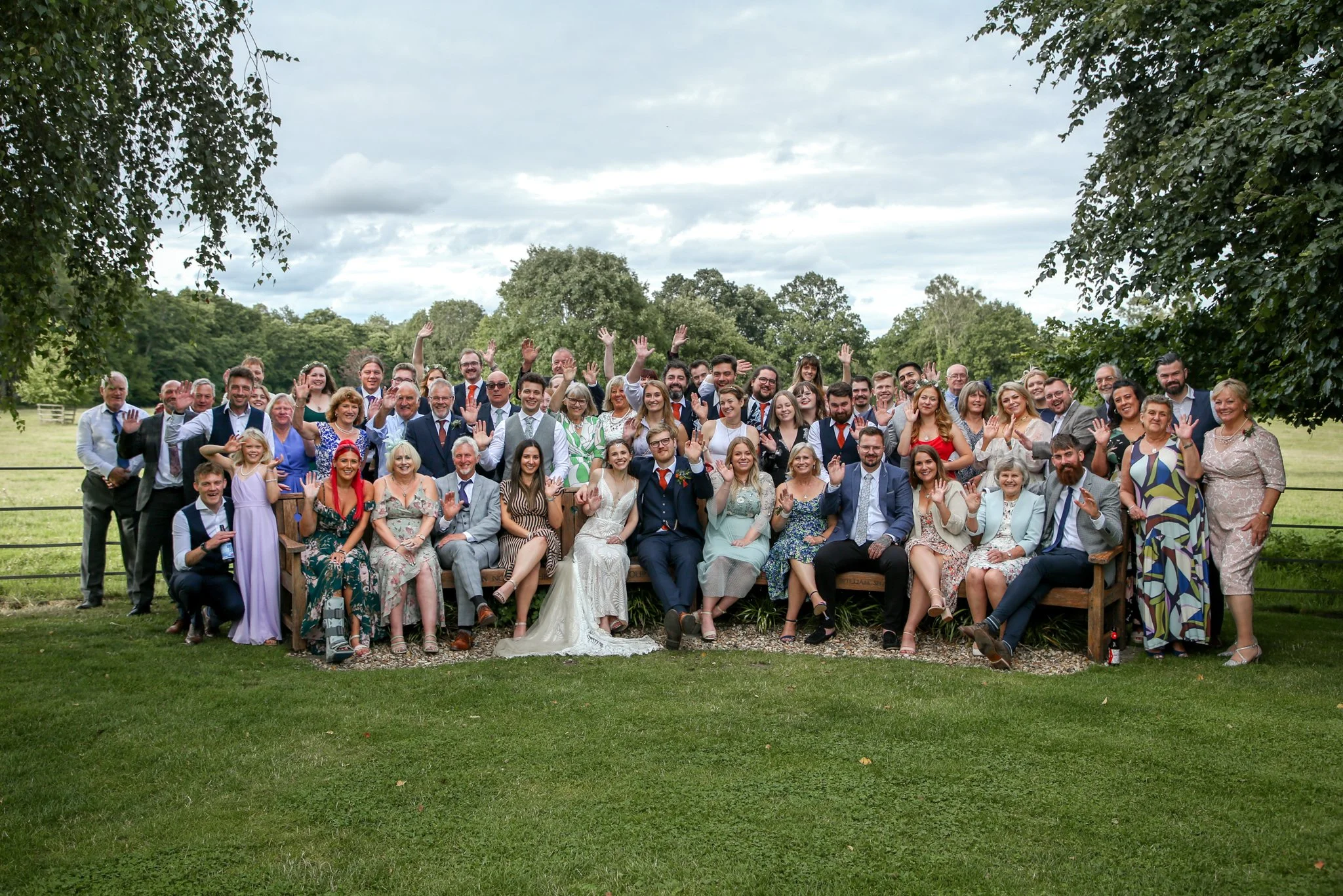 Large group of people gathered outdoors in a park for a celebration, with many waving and smiling at the camera.