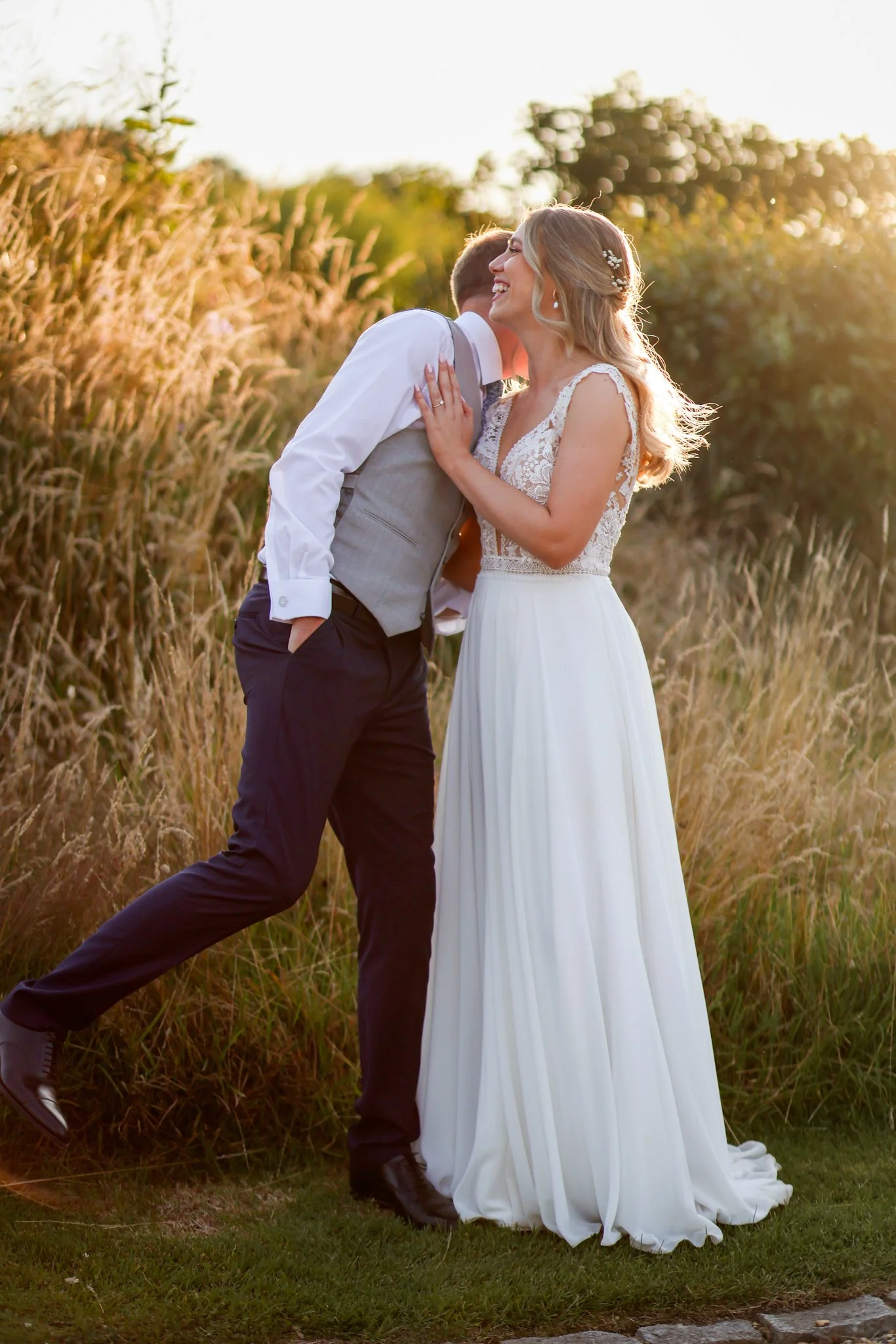 A bride and groom smiling and hugging outdoors at sunset, with tall grass and trees in the background.