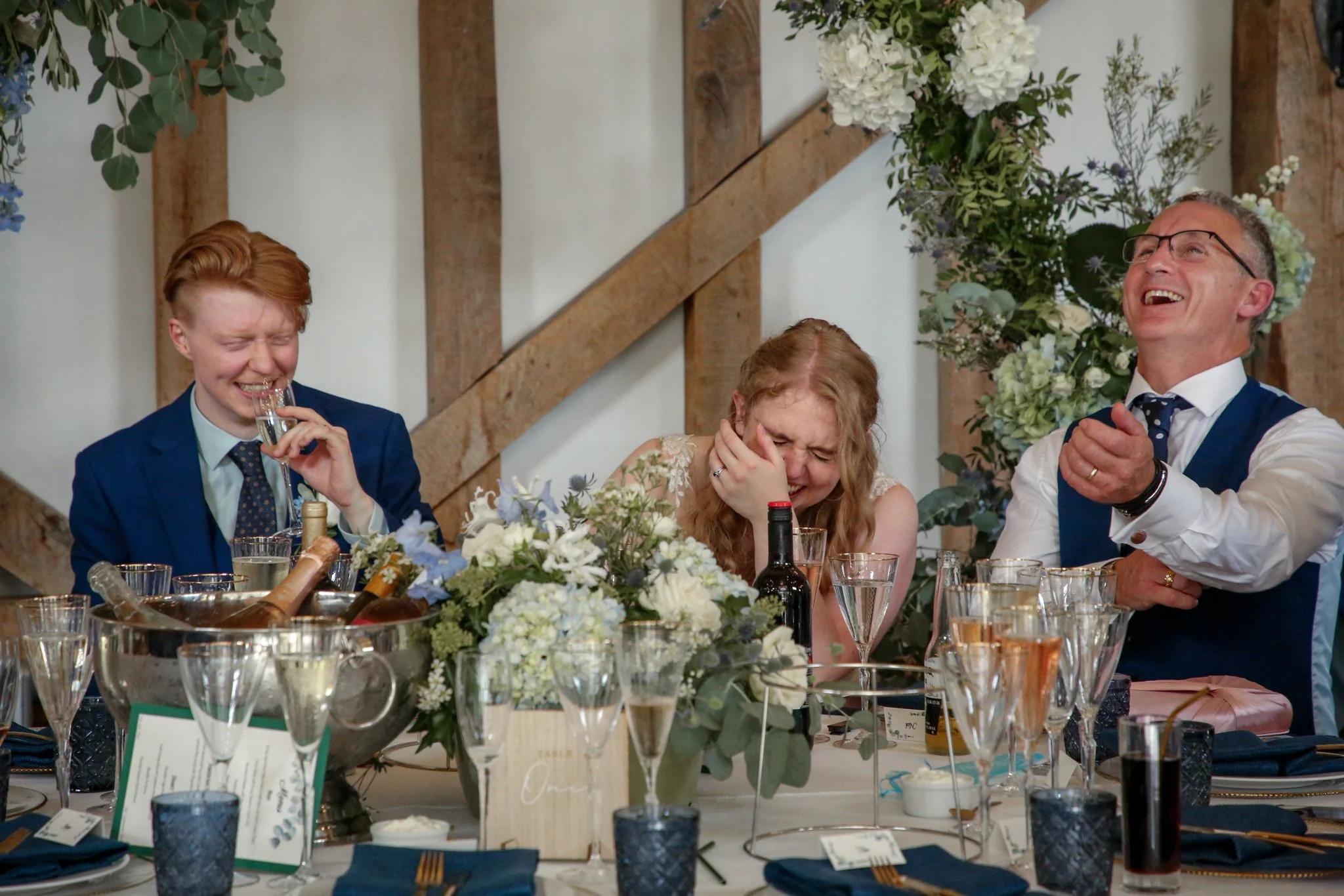 People at a wedding reception sitting at a decorated table, laughing and enjoying drinks, with floral centerpieces and place settings.