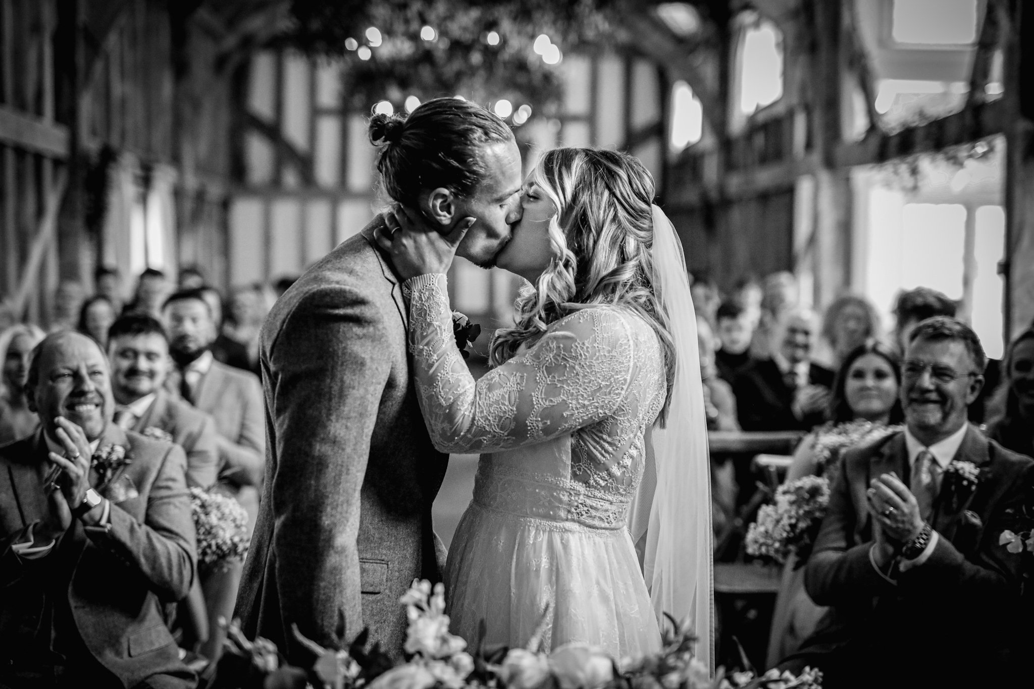 A bride and groom kiss during their wedding ceremony inside a rustic wooden venue, with guests watching and smiling.