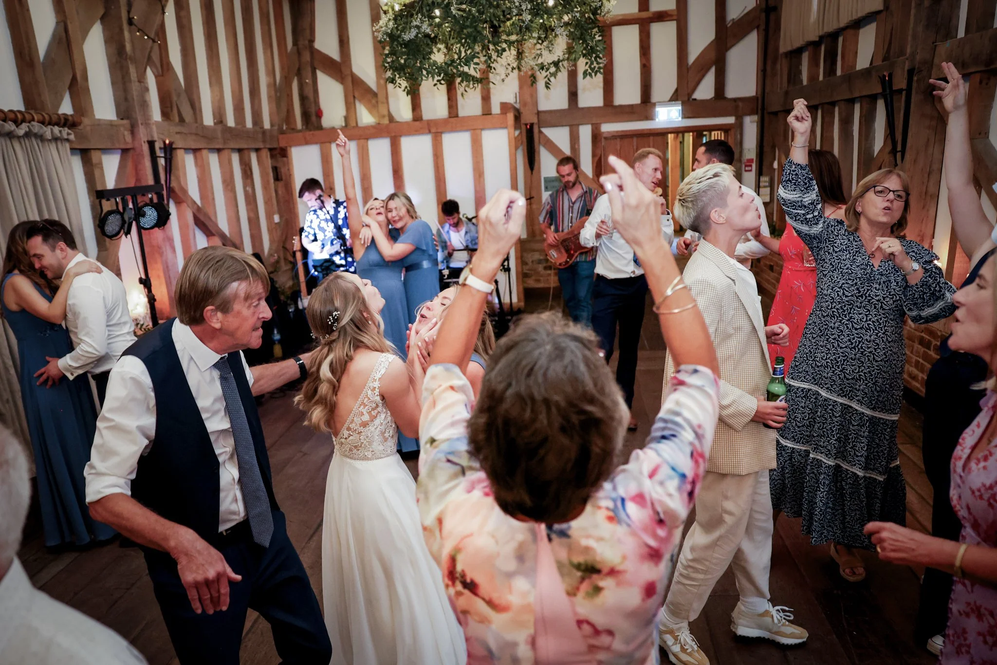 People dancing and celebrating at an indoor event with wooden walls, some are embracing and smiling, dressed in semi-formal attire.