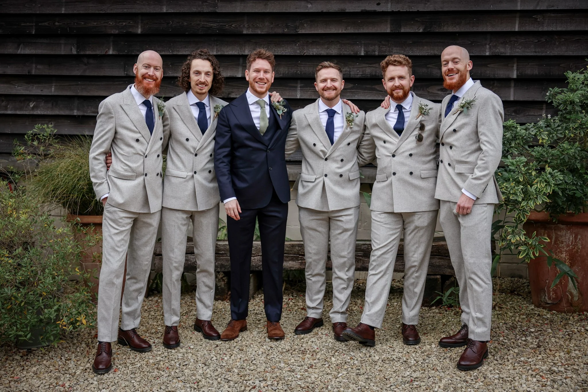 Six men dressed in suits, standing outdoors in front of a wooden wall, smiling and posing for a photo, with some men having beards and one with curly hair, and plants on both sides.