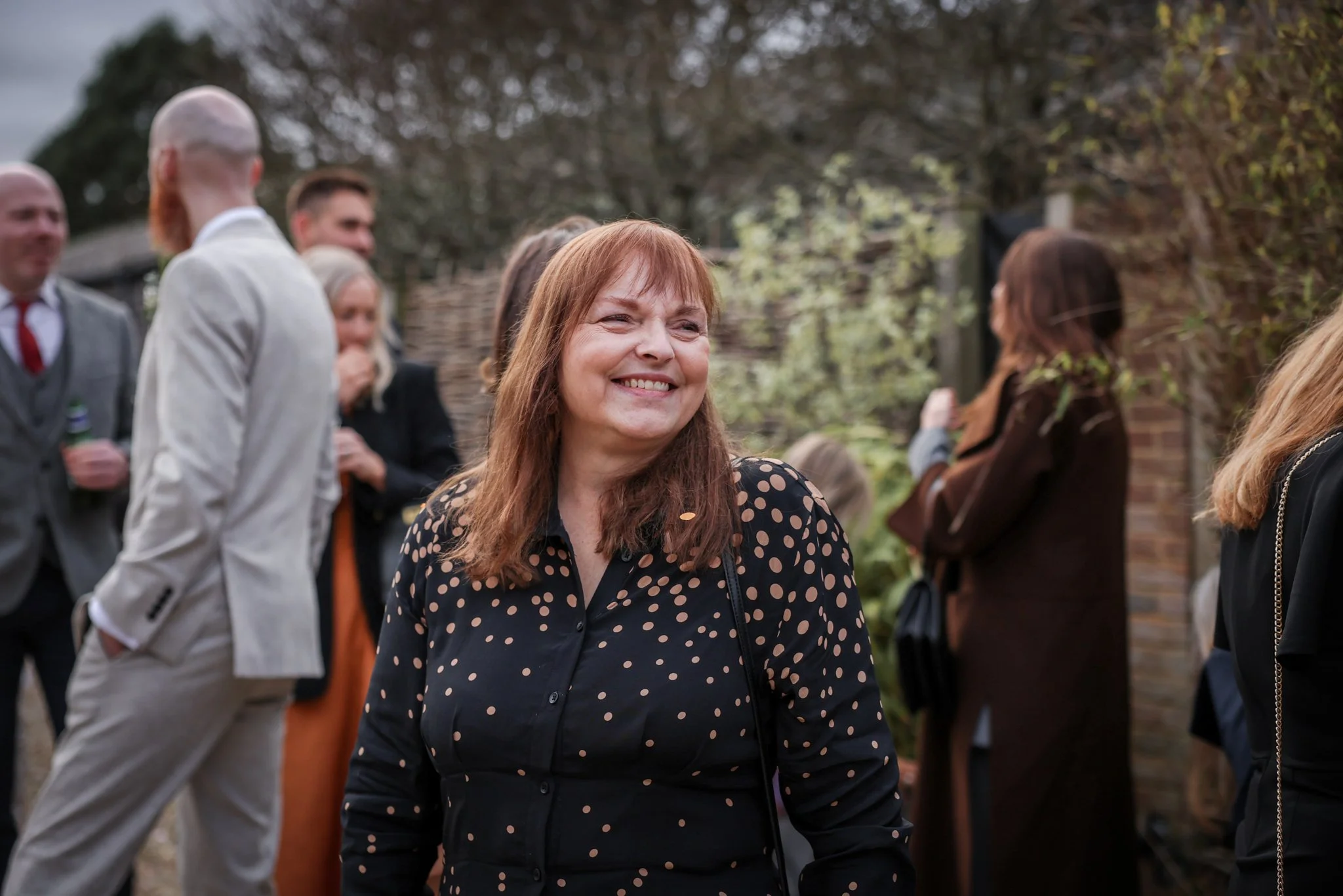 A woman with long red hair smiling in front of a group of people outdoors on a cloudy day.