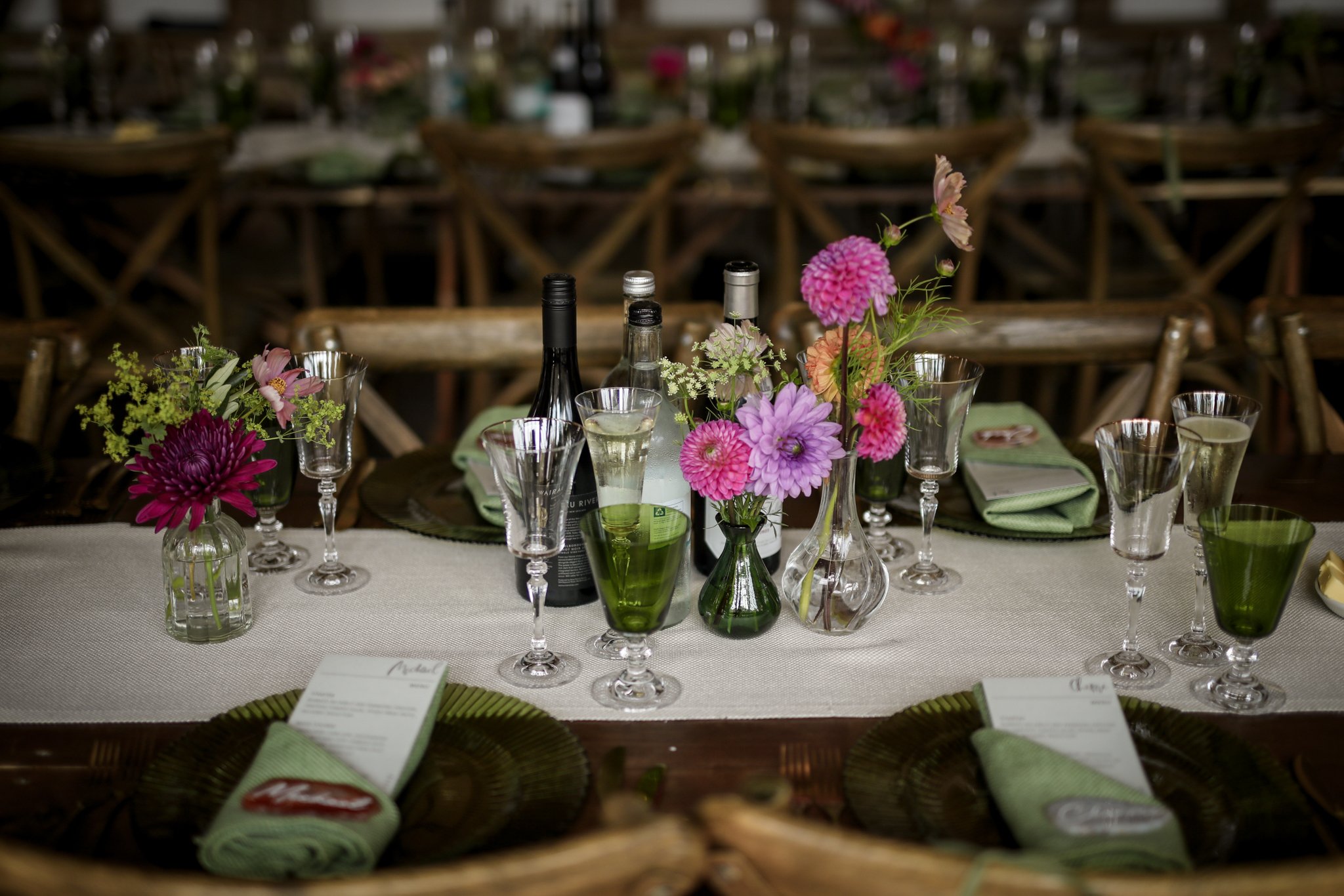 A table set for an event with vases of pink and purple flowers, wine bottles, glasses, and green napkins.