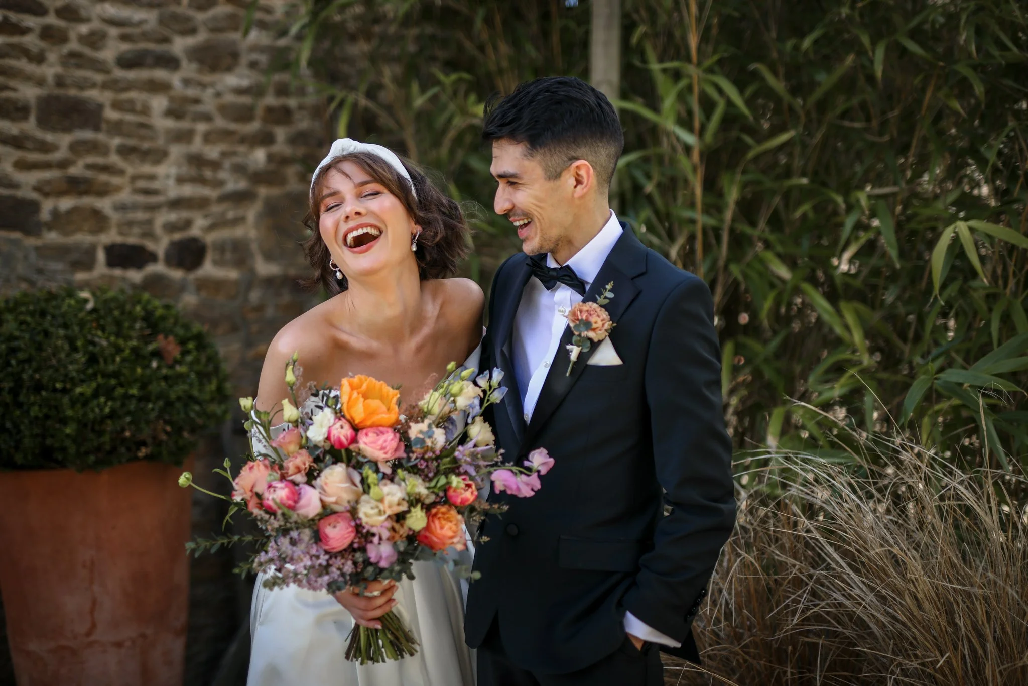 A bride and groom laughing together outdoors, with the bride holding a large bouquet of colorful flowers, in front of a stone wall and greenery.