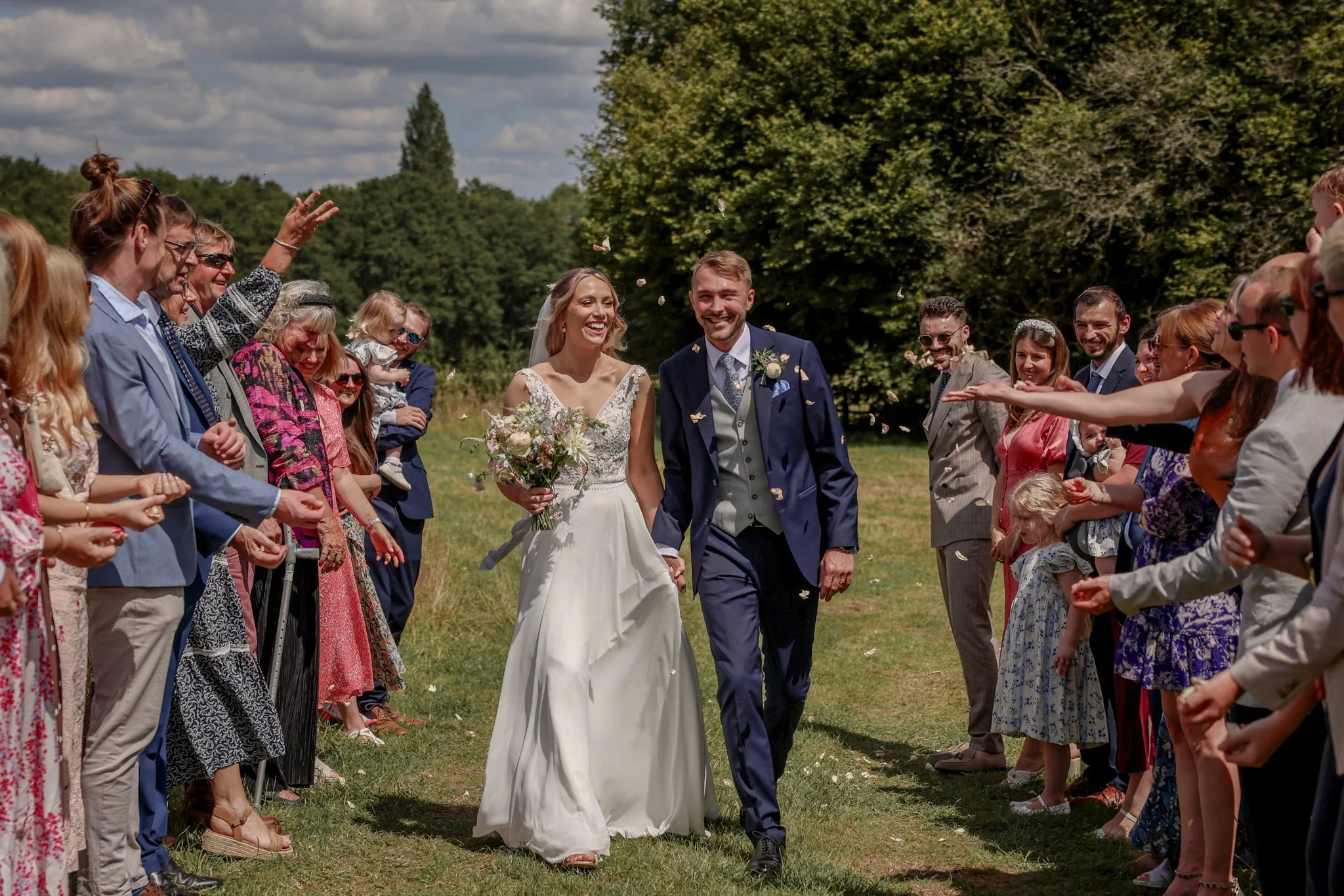 A bride and groom walking arm in arm through a crowd of guests at an outdoor wedding ceremony, smiling and throwing flower petals in the air. The guests are standing on both sides, dressed in colorful attire, beneath a partly cloudy sky with green tr