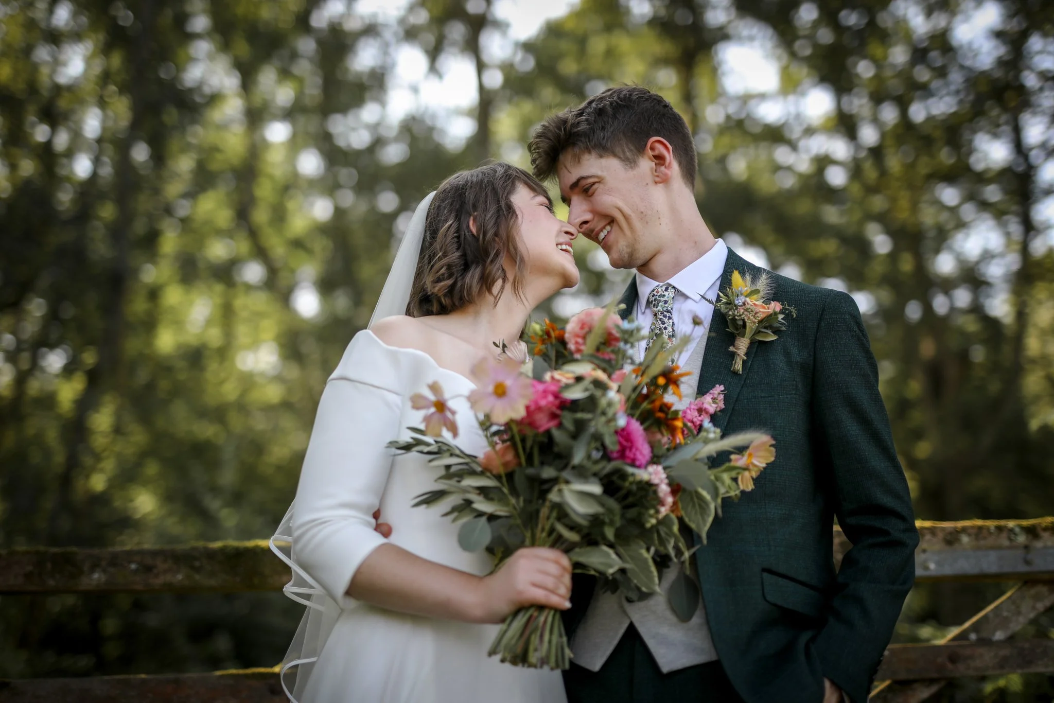 A bride and groom smiling and touching foreheads outdoors with trees in the background. The bride is holding a large colorful bouquet of flowers.